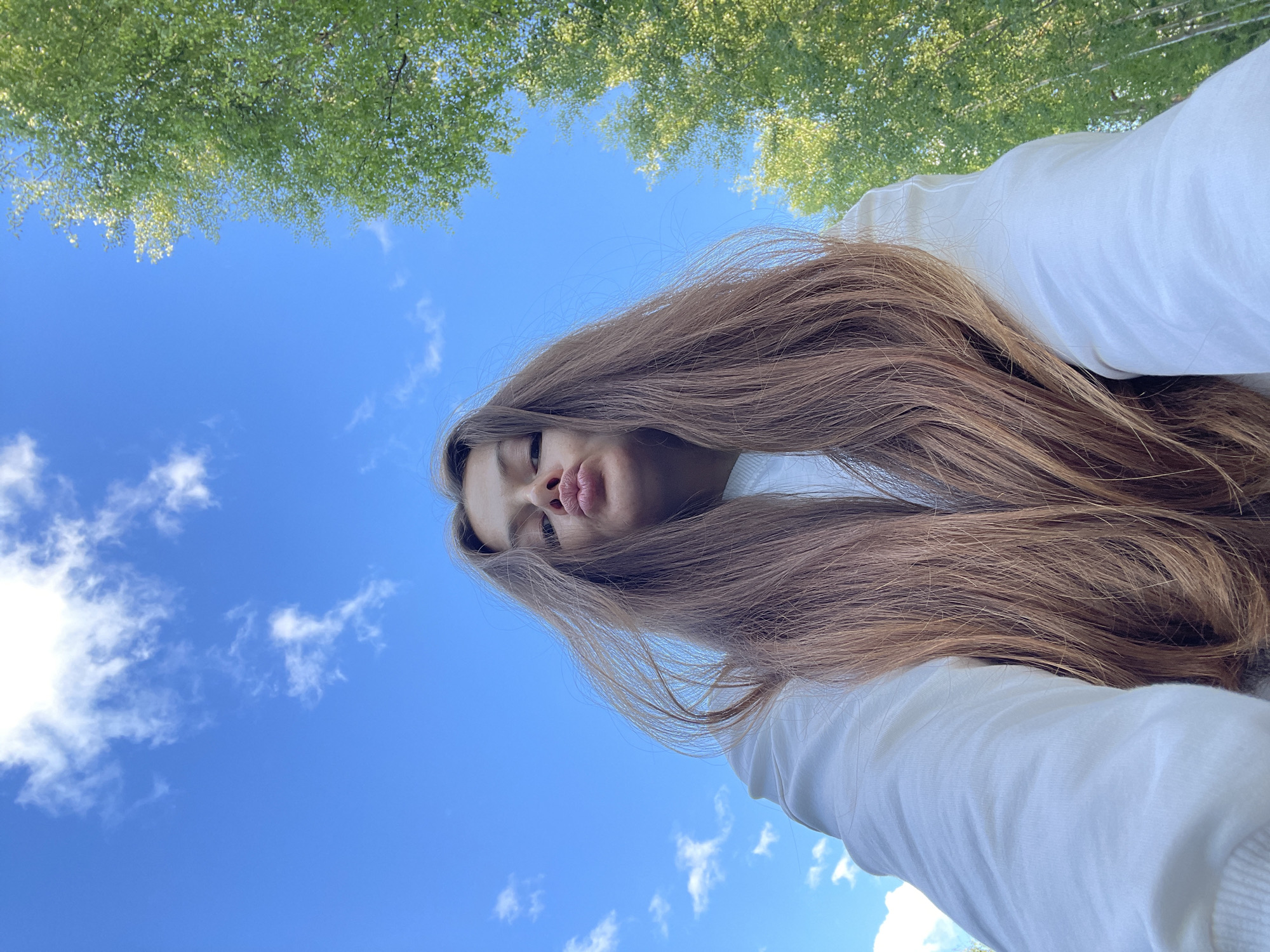 The image shows a young woman with long, light brown hair, looking upwards at the sky. She is wearing a white sweatshirt. Her mouth is pursed in a kissing gesture. The background consists of a bright blue sky with some scattered white clouds and the green foliage of trees. The perspective is from below, looking up at the woman. The scene appears to be outdoors, possibly in a park or a wooded area. There is no indication of the specific location in the city of Заречный, Russia.