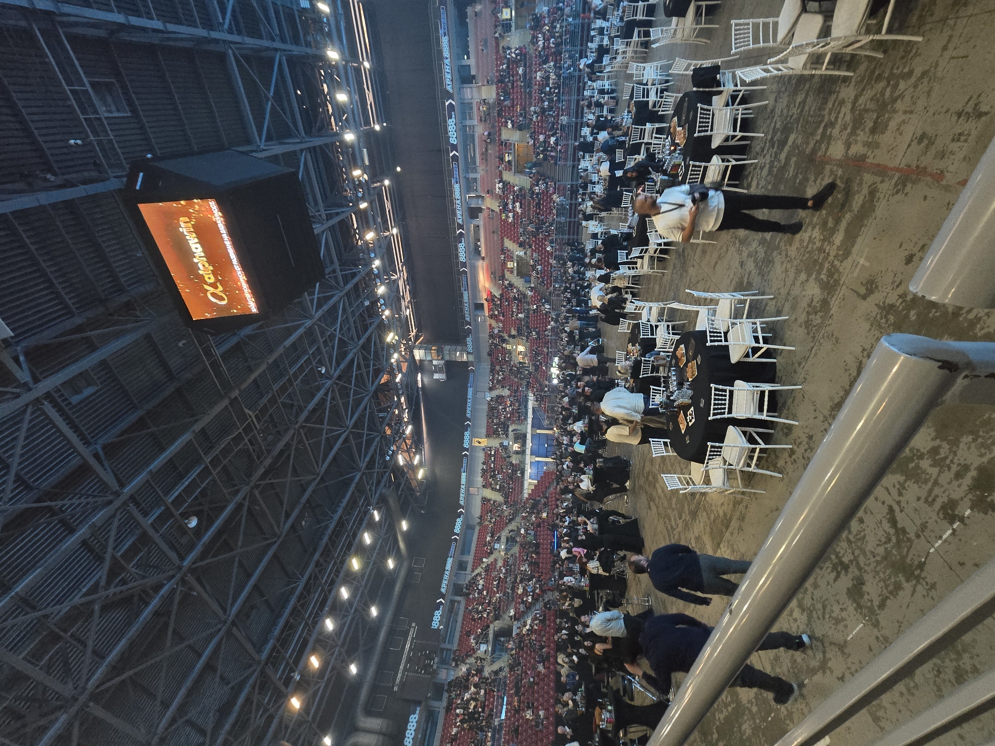 The image depicts a large indoor event taking place in Arena 8888 in Sofia, Bulgaria, as clearly indicated by the repeated text "АРЕНА 8888 СОФИЯ" on display screens. The setting suggests an evening or nighttime event, lit by numerous artificial lights mounted on the dark, industrial-style ceiling trusses.
The venue's floor is set up with numerous circular tables covered with black tablecloths and surrounded by white chairs. Many people are visible, some seated at these tables, while others stand and move between them, indicating mingling or a formal dinner setup. Beyond the floor area, tiered stadium seating, predominantly red, rises, also occupied by attendees.
A large overhead screen displays the text "alphawin" and "Celebrating 20 years," suggesting the event is an anniversary celebration sponsored by Alphawin. Additional screens and banners around the arena's perimeter also display "8888" and "АРЕНА 8888". The perspective is from an elevated position, overlooking the event space, with a gray railing visible in the foreground. The overall atmosphere appears to be a large-scale corporate event, gala, or special gathering within a major arena.