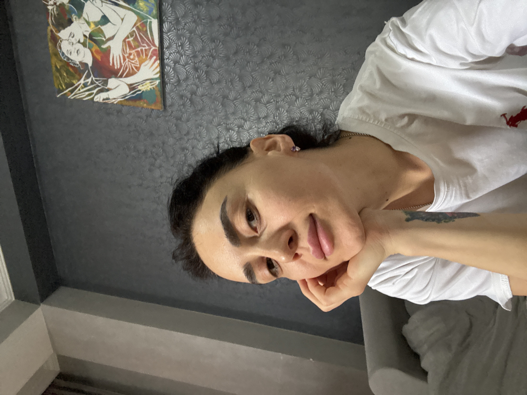 The image is a selfie of a person with dark hair and light skin wearing a white t-shirt. They are resting their chin on their hand, which has a tattoo on the wrist. The background consists of a patterned grey wall with a framed painting hanging on it. The painting appears to depict two figures in an embrace. The bottom left corner of the frame is visible. The person is sitting in a grey chair. The exact location within Tbilisi, Georgia, is not discernible from the image.