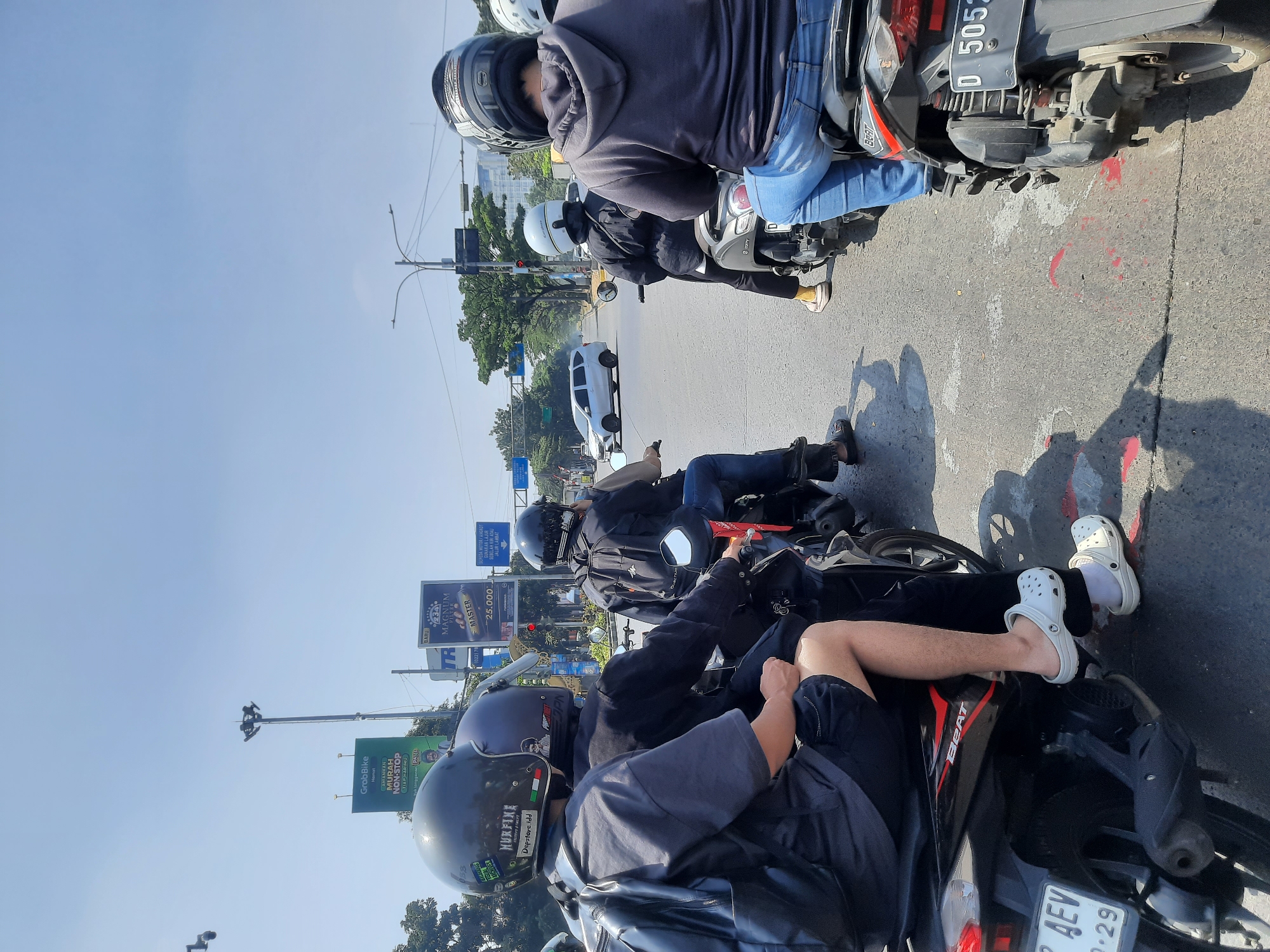 The image captures a street scene in Bandung City, Indonesia, likely during daylight. Several individuals are riding motorcycles in what appears to be traffic. In the foreground, two people are on a black motorcycle, with the person in the back wearing a black backpack and a grey t-shirt, and the person in the front wearing a black helmet with stickers. They are both wearing casual clothing, including shorts and white Crocs. To their right, another motorcycle with two riders is visible. The rider in front is wearing a black helmet and jacket, and the passenger behind is wearing a blue hoodie and jeans. Further ahead, a white car is driving through an intersection. The environment includes traffic lights, street signs, billboards, and trees, suggesting an urban setting. The road surface is asphalt, with some red markings on the ground. The sky is clear and blue.