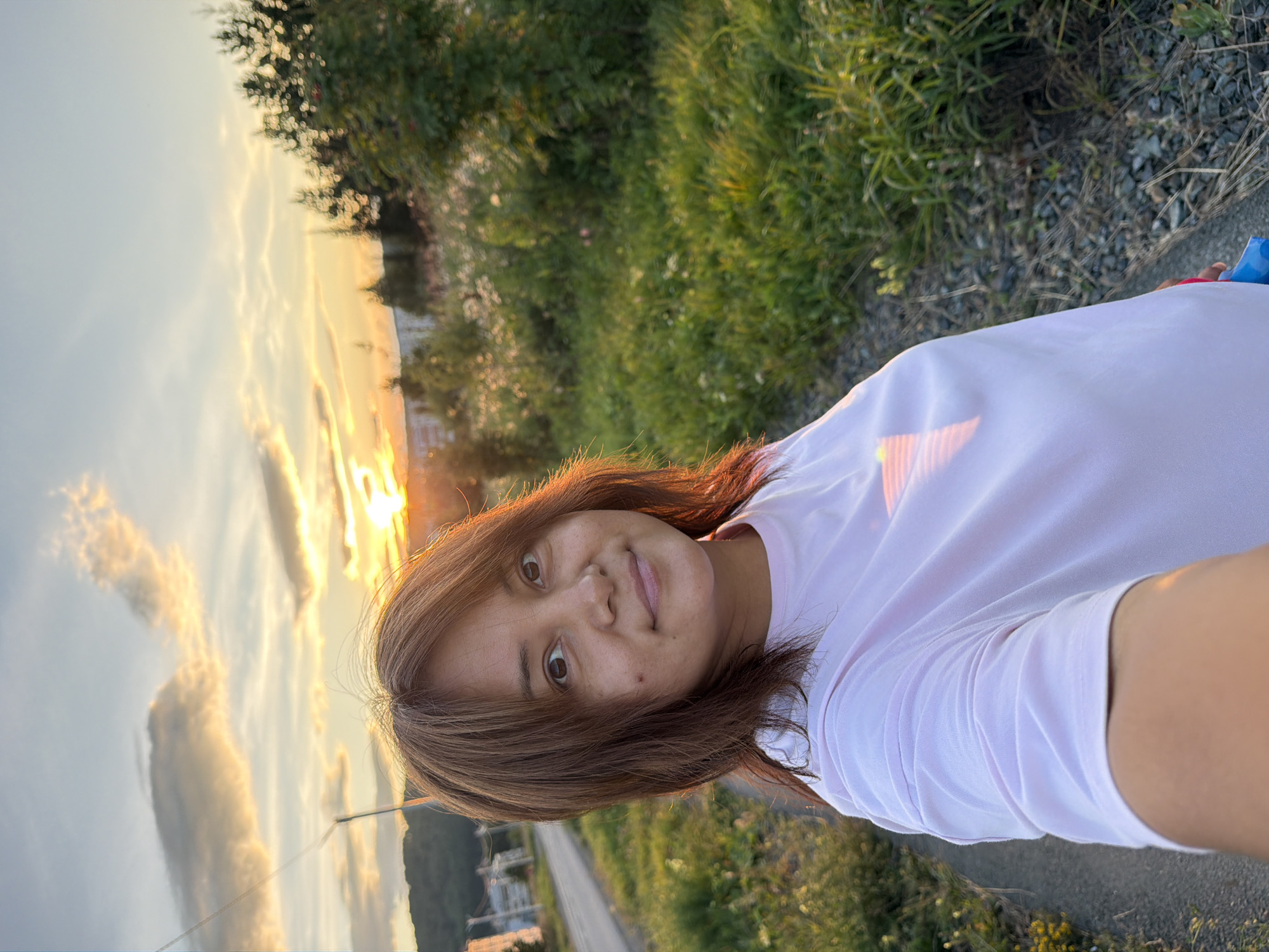 A medium shot selfie captures a woman with brown hair and fair skin looking towards the camera. She is wearing a pale pink t-shirt and is positioned in the foreground. The background reveals a sunlit landscape with a road on the left and a grassy area with trees and some buildings in the distance on the right. The sun is setting, casting warm light and creating a sunset effect in the sky, with some clouds present.