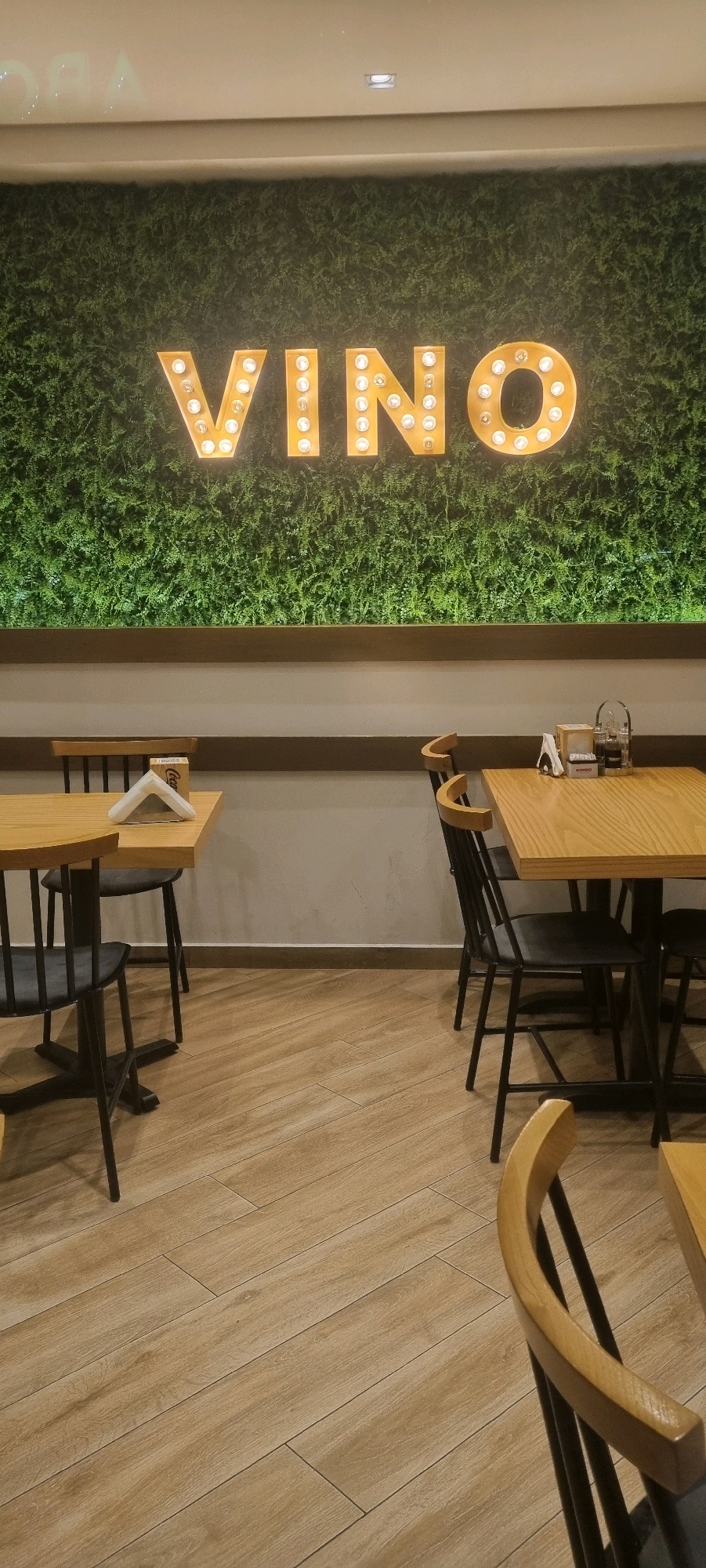 An interior scene of a restaurant or cafe is depicted, likely in Bashkia Durrës, Albania, based on the signage and ambiance. The dominant feature is a lush green wall, possibly artificial foliage, serving as a backdrop. Prominently displayed on this wall is a large, illuminated sign spelling out "VINO" in vintage-style marquee letters with exposed bulbs. Below the green wall, a thin, dark wooden shelf runs horizontally.
In the foreground and mid-ground, several wooden tables with dark chairs are arranged. One table in the left foreground has a white napkin holder with napkins and a Coca-Cola branded box. Another table in the mid-right has a similar setup, including a napkin holder and what appears to be a condiment caddy with bottles. The floor is covered with light-colored, wood-look tiles laid in a staggered pattern. The overall lighting is warm and inviting, suggesting an evening or dimly lit interior. No people are visible in the frame.