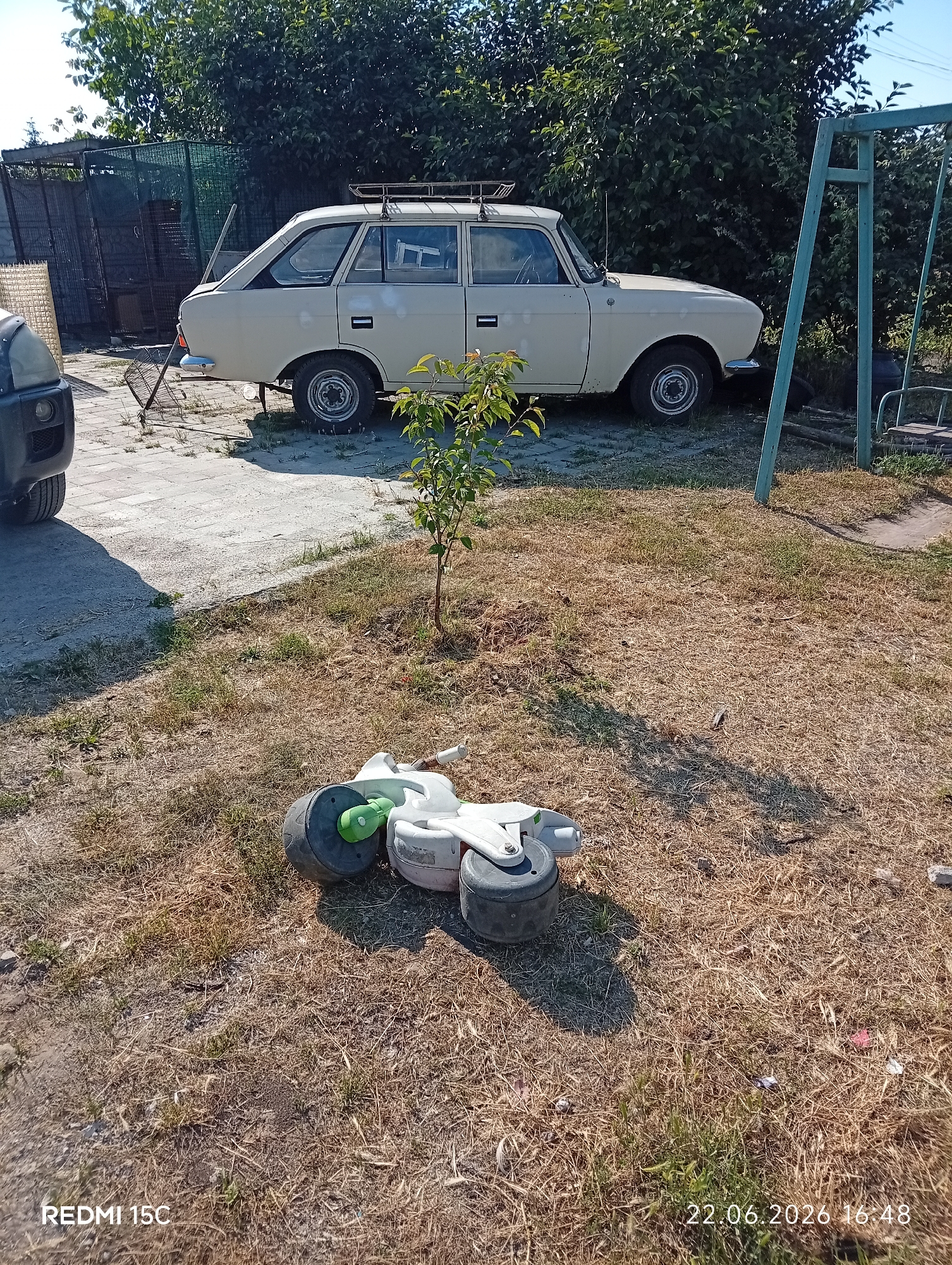 A cream-colored station wagon is parked on a paved surface in a yard. A portion of a black vehicle is visible on the left. In the foreground, a white toy bicycle with green and black wheels lies on the dry grass. A small tree is positioned between the car and the toy. In the background, there are trees, a metal fence, and a green swing set. The sky is clear and blue. The overall scene suggests an outdoor setting, possibly a residential backyard. The timestamp "22.06.2026 16:48" and "REDMI 15C" are visible in the bottom right and left corners of the image, respectively.