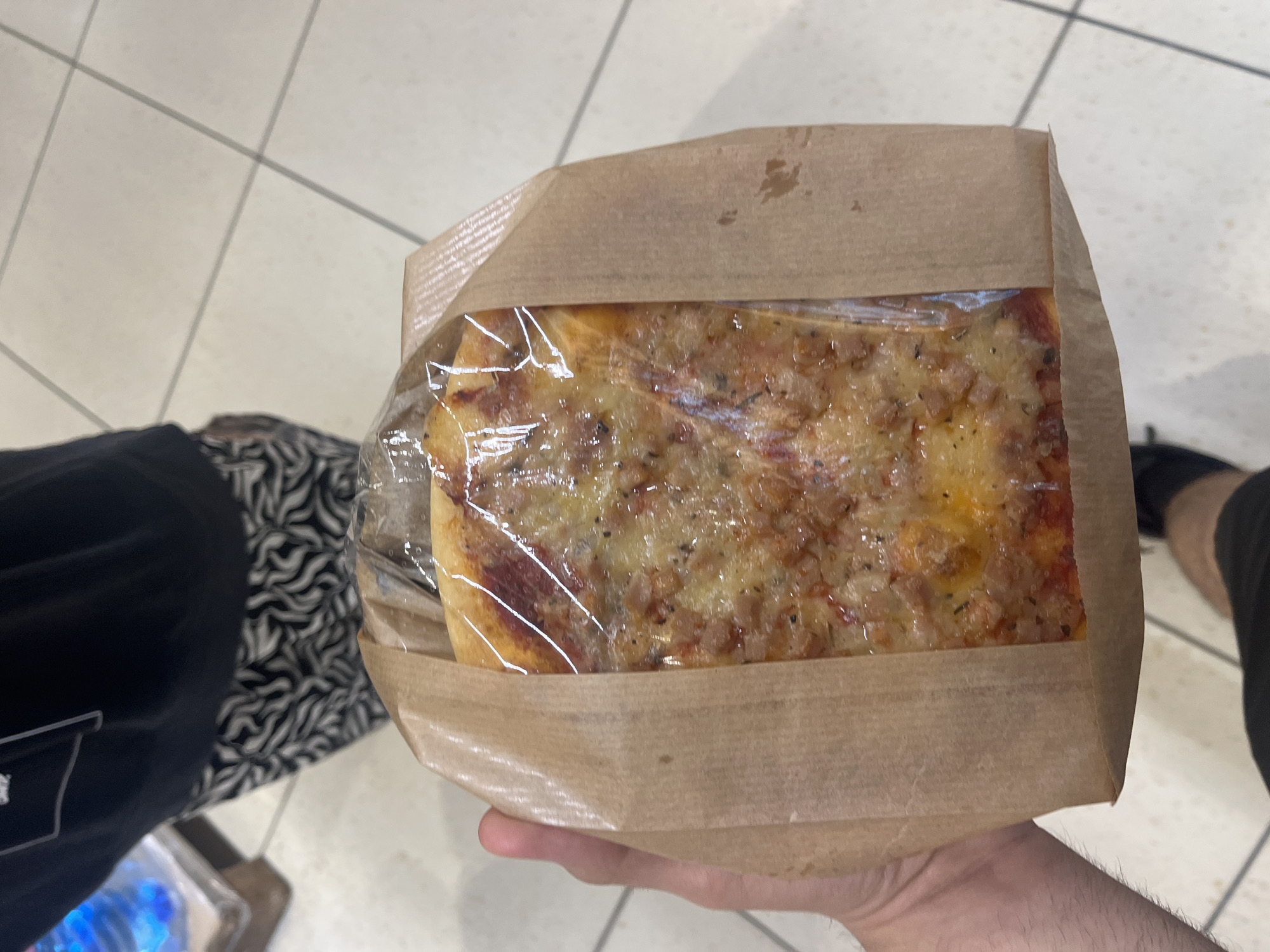A person is holding a square slice of pizza in a brown paper bag, covered in clear plastic wrap. The pizza has a visible topping of cheese and sausage. The person is standing indoors on a tiled floor. In the background, parts of the person's clothing are visible: patterned pants and a dark t-shirt. Another person is partially visible at the right side of the image with dark athletic pants and black shoes. The location is not discernible as Blagoevgrad, Bulgaria, without additional information.