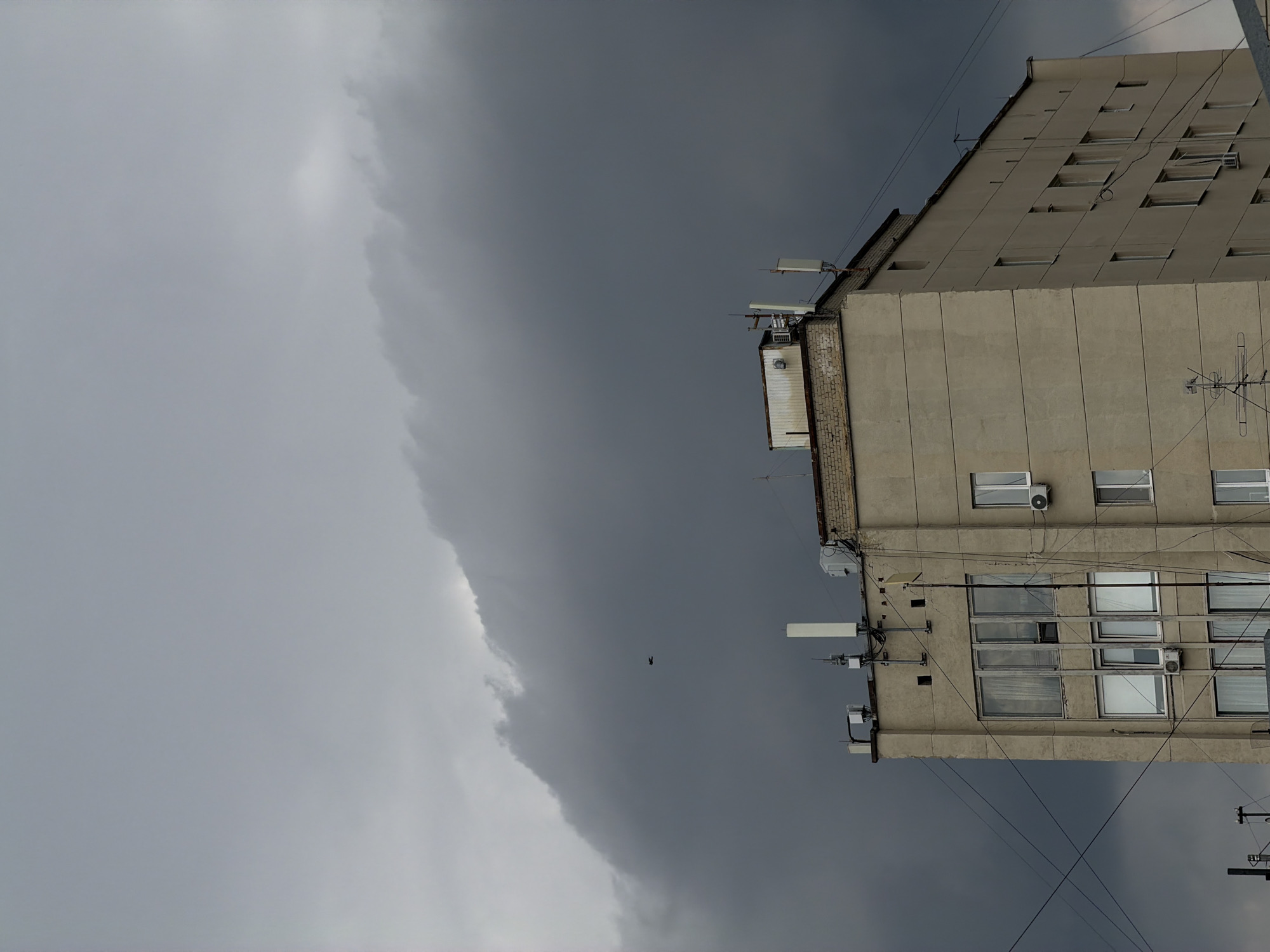 This is a medium-shot, eye-level, natural light photograph taken from a low angle of a concrete building with a light beige facade against a dark, ominous sky. The building has multiple windows, some with curtains visible inside. The top of the building is adorned with various antennas and equipment, suggesting it is a commercial or telecommunications facility. Several power lines crisscross the image in the foreground. In the distance, a single bird is visible against the heavy, gray clouds. The scene conveys a sense of impending weather. The building is located in the city of Saratov, Russia.