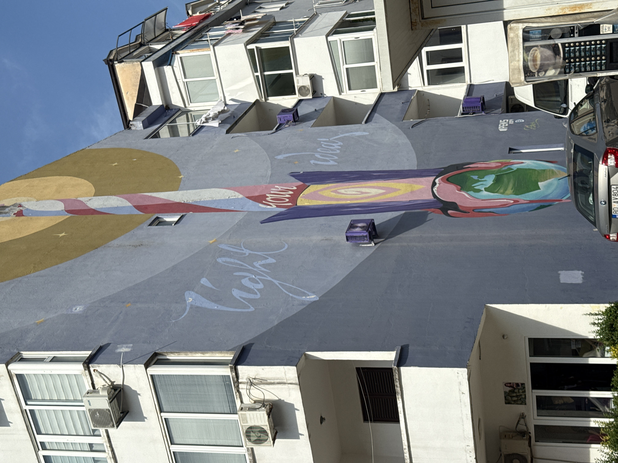 The image depicts a mural on the side of a building in Budva, Montenegro. The mural features a tall, swirling object that resembles a rocket or a stylized flame, painted in red, white, and yellow, emerging from an abstract representation of the Earth. The words "light your ideas" are written in flowing blue script across the mural. The building itself is painted a muted gray and has several balconies with white railings, typical of apartment buildings in the area. There are air conditioning units mounted on the exterior walls. To the right of the mural, another building is visible, also featuring balconies and windows. In the foreground, a silver car is parked, and a coffee vending machine is positioned on the right side of the frame. The sky is clear and blue. The overall scene suggests an urban environment with a focus on public art.
