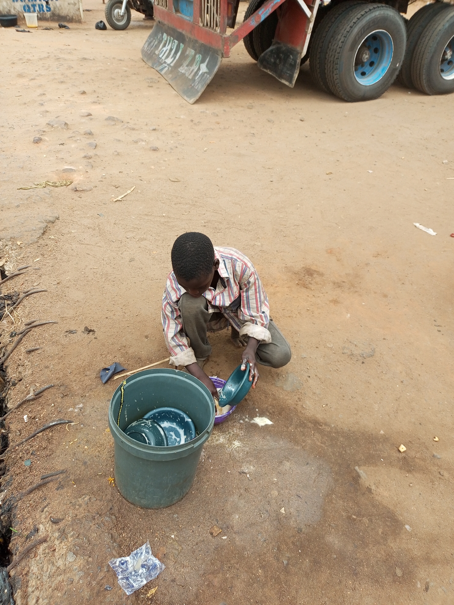 In Gombe, Nigeria, a young boy crouches on a dirt surface, engaged in washing dishes. He wears a checkered shirt and dark trousers. To his left, a large, dark green bucket is filled with water and several blue and green plates. He holds a smaller blue bowl in his hands, pouring water from it. Nearby, a dark blue item, possibly a sponge or brush, lies on the ground. The background shows the tires of a large truck, and a partially visible sign with text. The environment is an outdoor, unpaved area.