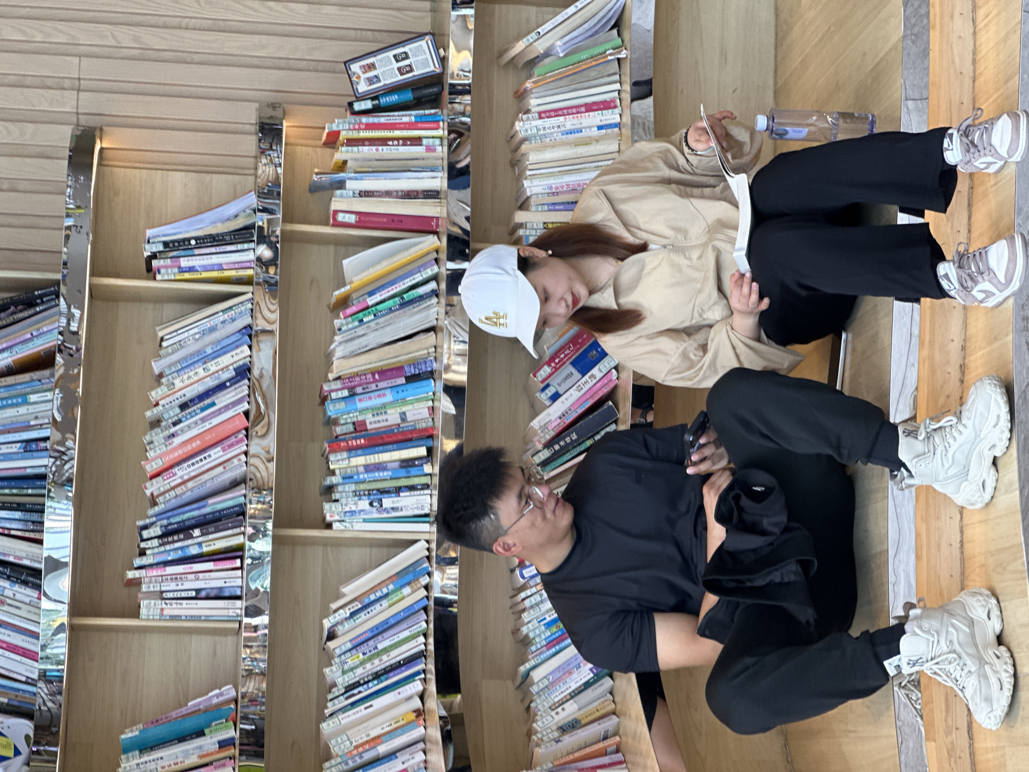 A high-angle shot of a reading area, perhaps in a bookstore or library, in Liangjiang, China. The photograph is rotated 90 degrees counter-clockwise. A shelf filled with numerous books takes up a significant portion of the left side of the image. Two people are sitting on the floor in front of the shelf. A young woman is wearing a tan jacket, black pants, and white shoes. She has brown hair and is wearing a white baseball cap with gold lettering. She holds an open book in her hands and looks down at it. There is also a water bottle next to her. A young man, wearing black-rimmed glasses and dressed in a black t-shirt and black pants, sits beside her. He looks to his left, away from the woman. Both individuals are sitting on what appears to be wooden flooring. The overall environment is well-lit.