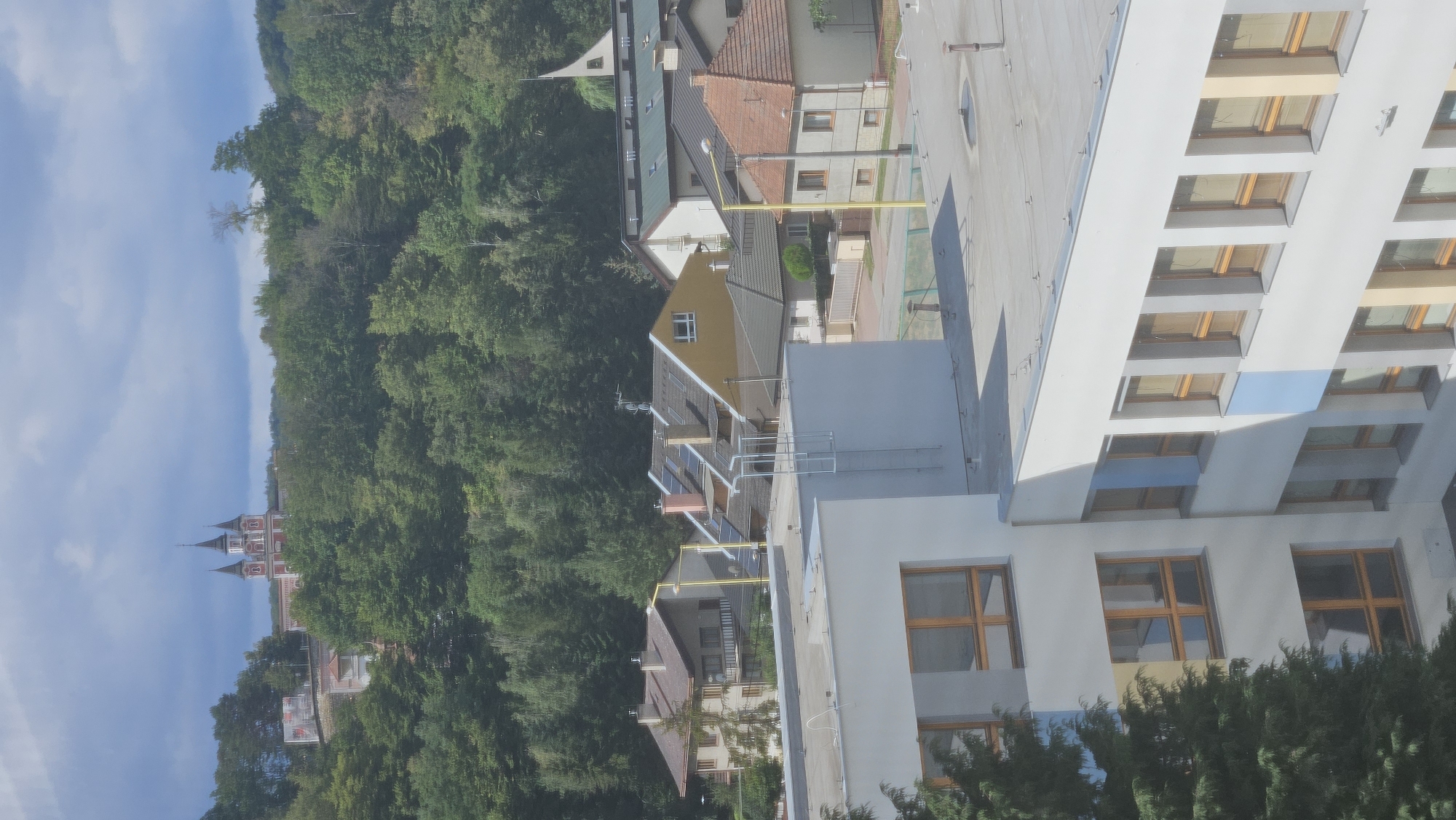 The image shows a view of a town with a distinctive red and white building with multiple spires on a hill in the background, surrounded by trees. In the foreground, there are several residential buildings and a modern white building with many windows. The sky is partly cloudy. The red and white building in the background is the Orthodox Church of St. Nicholas in Prešov, Slovakia.