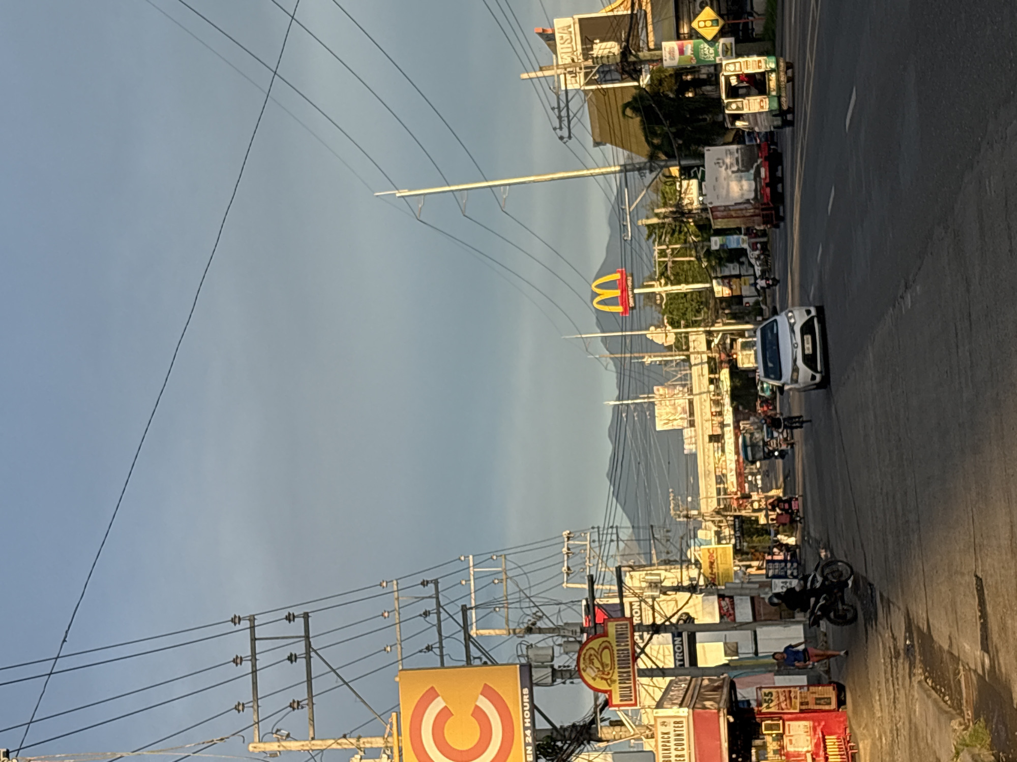 The image depicts a street scene in Davao City, Philippines, likely in the early morning or late afternoon given the warm, low-angle sunlight. The road is paved and appears to be a busy thoroughfare with multiple lanes of traffic. Several vehicles are visible, including cars, a motorcycle with a rider, a truck, and a three-wheeled vehicle. Pedestrians are also present on the sidewalk.
Along the sides of the road, numerous commercial establishments are visible, indicated by signs and awnings. Prominent among these are a McDonald's restaurant, a "Bulk Pack Burger Counter," and other businesses with signage that is partially obscured or difficult to read. Overhead, a complex network of electrical wires and utility poles crisscrosses the sky, characteristic of urban infrastructure. In the background, a range of mountains is visible against the sky, providing a natural backdrop to the urban environment. The overall atmosphere suggests a vibrant and active commercial district within Davao City.