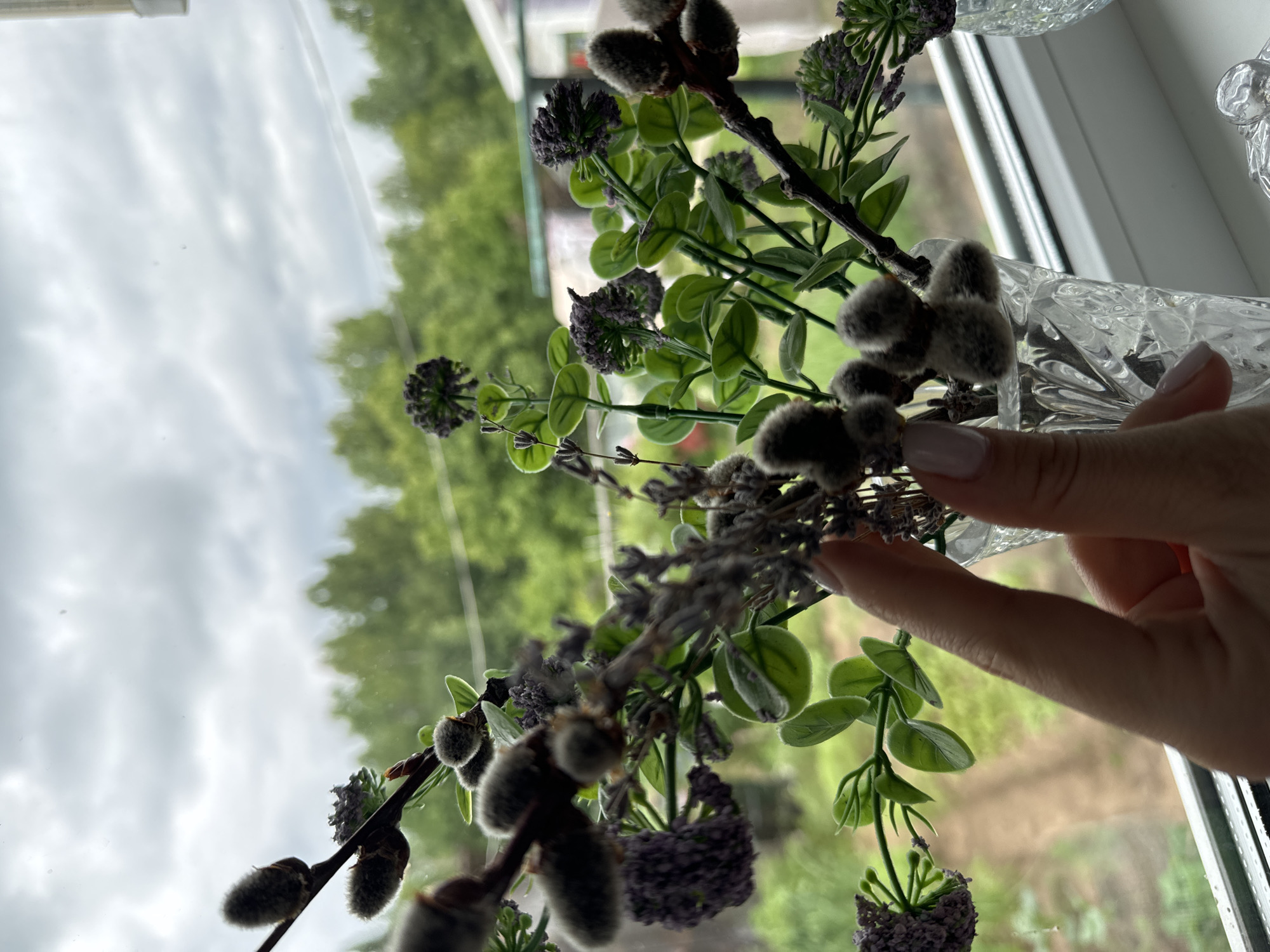 An outdoor scene viewed through a window shows a close-up of a bouquet of artificial flowers, including pussy willow branches and small purple florets, arranged in a clear glass vase. A person's hand with light-colored nail polish is visible, gently touching one of the pussy willow branches. The bouquet is placed on a white windowsill. Beyond the window, a blurred landscape of green trees and a cloudy sky is visible. The setting appears to be the city of Ponaryrevo, Russia, given the context provided.