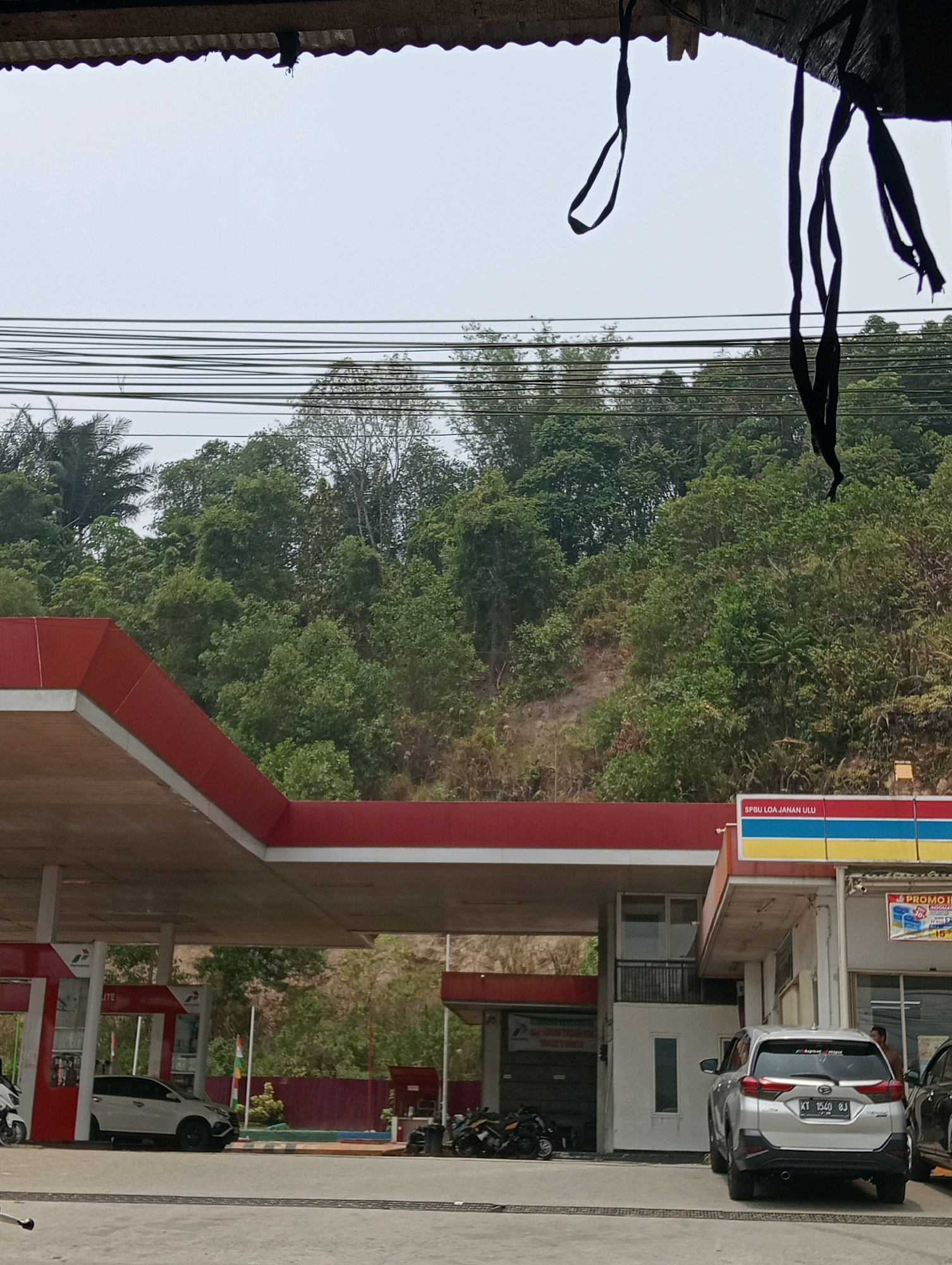 The image captures a view of a gas station in Loa Janan Ulu, Indonesia. A red and white canopy extends over several parking spots. A silver SUV with license plate KT 1540 BJ is parked under the canopy, facing away from the viewer. To its left, a white hatchback is parked under the same canopy. A person can be seen standing near the entrance of a building to the right of the SUV. Several motorcycles are parked to the left of the white hatchback and further to the left. The background is dominated by a lush, green hillside covered in trees. Utility wires are strung across the sky. The sky is overcast.