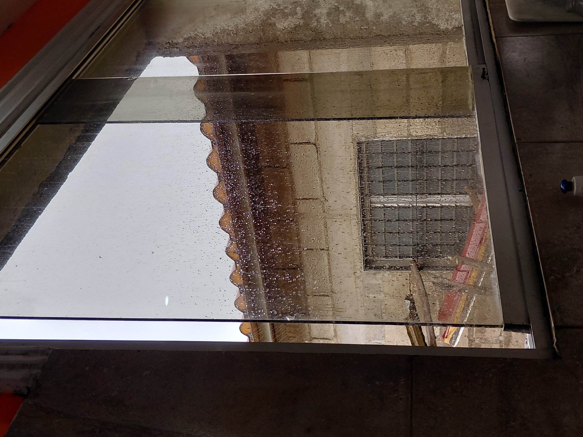 The image displays an outdoor scene viewed through a wet, reflective glass surface, likely a window or skylight, from an interior or covered vantage point. Numerous small water droplets cover the glass, indicating rain or recent precipitation.
Reflected in the glass is a building featuring a light-colored, possibly concrete or stucco, wall. A window with a distinctive dark grid pattern, suggesting either bars or small panes, is clearly visible within this wall. Above and to the left of the window's reflection, a section of a reddish-brown, possibly terracotta, roof with a wavy, corrugated edge is reflected. Faint reddish and yellow colors are also reflected below the window, hinting at other architectural elements or coverings.
The transparent surface is framed by a metallic, silver-colored border. In the immediate foreground, dark, possibly ceramic, tiled flooring is visible on the bottom right and left sides, establishing the indoor or sheltered nature of the viewing position. A small white and blue object, possibly a bottle cap, rests on the tiled floor in the far right corner. An orange-colored wall or structure is visible in the upper left section of the overall frame, beyond the glass.
The scene appears to be set during the daytime, likely under overcast skies due to the rain. This perspective of local architecture is situated in Santa Ana, Venezuela. No visible text is present in the image.