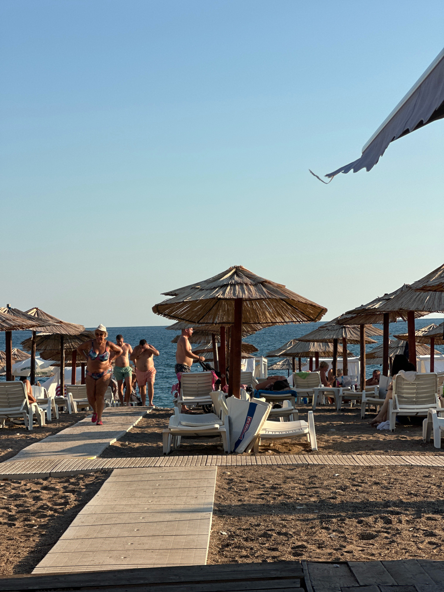 This image captures a sun-drenched beach scene in Boreti, Montenegro. The foreground is dominated by a wooden boardwalk, leading towards the sea. Sandy ground, dotted with small pebbles, surrounds the boardwalk. Several thatched umbrellas are erected on the beach, providing shade for rows of empty white lounge chairs.
In the midground, people are strolling along the boardwalk and on the sand. A woman in a colorful bikini walks towards the viewer, while several men in swim trunks walk away from the sea. Some individuals are seated on lounge chairs further back, enjoying the beach. The sea, a calm blue, stretches to the horizon, meeting a clear, pale blue sky. A dark awning extends from the upper right corner of the frame, partially obscuring the sky.