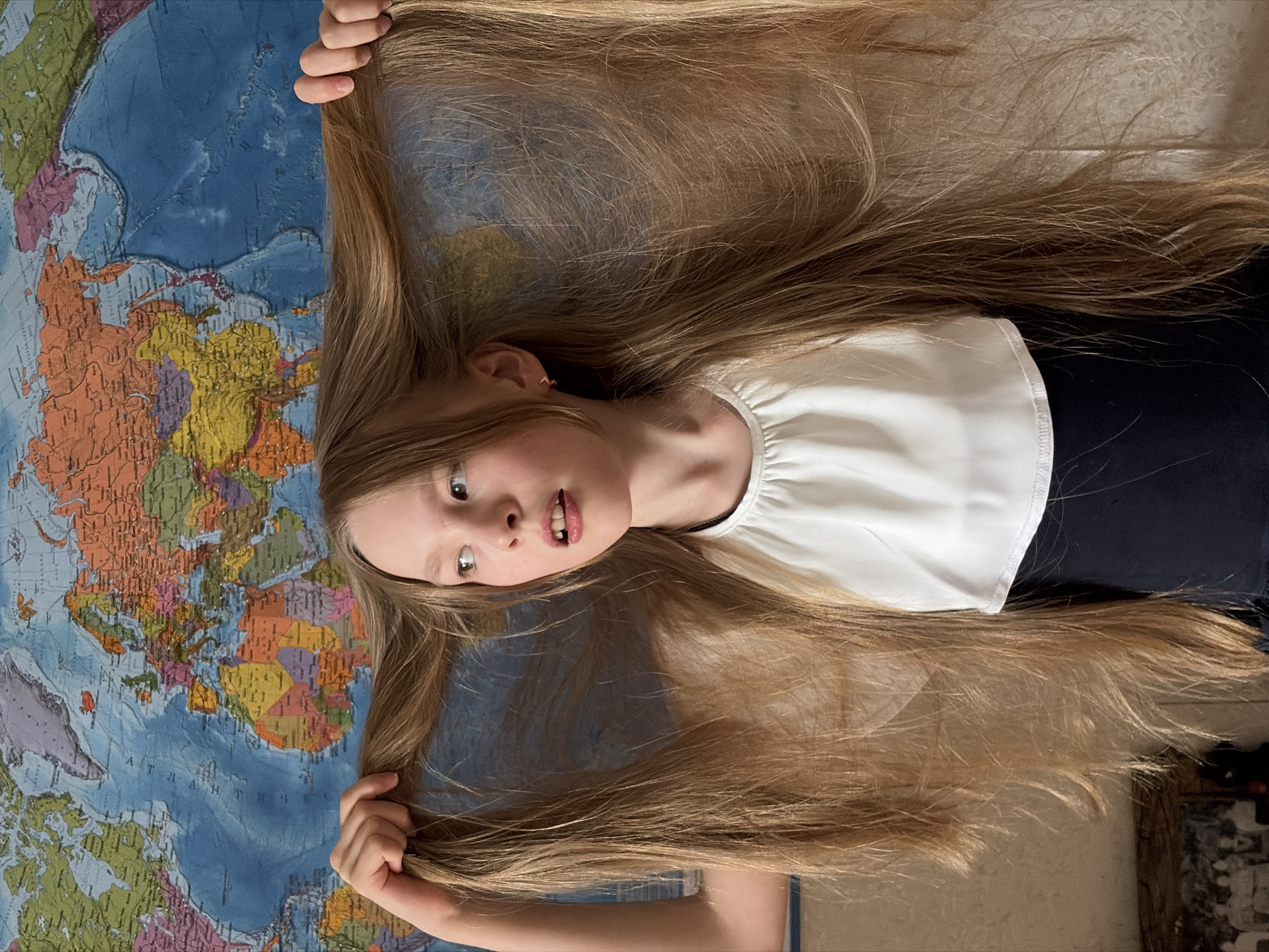 A young girl lies on her back on a world map. She is wearing a white blouse and a dark-colored bottom. Her long, light brown hair is spread out on either side of her head, held in her hands. The map is brightly colored with different countries delineated. The background is out of focus, showing part of a room with other objects. There is no indication of Izhevsk, Russia.