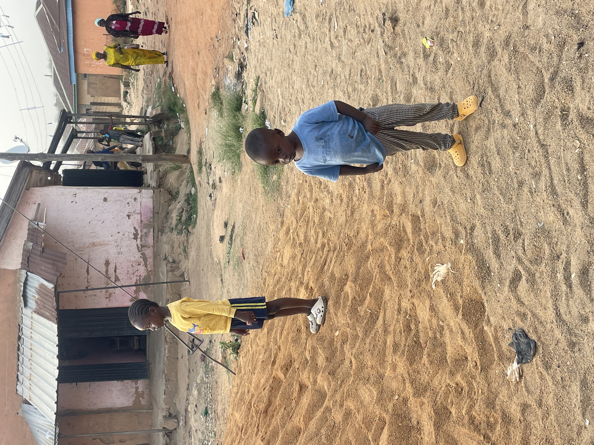 The image presents an outdoor scene in a residential area, characterized by sandy ground and simple architecture, consistent with the provided context of Jos, Nigeria.
In the foreground, two children are visible. To the right, a young boy with a short haircut wears a light blue t-shirt, black-and-white checkered pants, and yellow clog-style shoes. He looks towards the left with a calm expression. Further left, another child, possibly a girl, with braided hair, stands in a yellow long-sleeved shirt, dark blue shorts with yellow stripes, and white sneakers. This child also appears to be observing the surroundings. Both children are standing on the textured, light brown sand that covers much of the ground, interspersed with small tufts of dry grass and scattered pieces of litter, including plastic and dry leaves.
In the mid-ground and background, various structures and people can be seen. Two women, dressed in colorful traditional attire and head coverings, are walking along a dirt path. One wears a yellow outfit, and the other a darker patterned one. Behind them and to the left, several buildings are visible. These include structures with corrugated iron roofs, faded painted walls (one appearing pink), and some exposed brickwork. A wooden and metal framework, possibly supporting a makeshift shelter or market stall, is prominent on the left side, and a satellite dish is attached to one of the buildings.
The scene is illuminated by bright daylight, suggesting a clear day in either the morning or afternoon. The overall impression is that of everyday life in a local community. No discernible text is visible in the image.