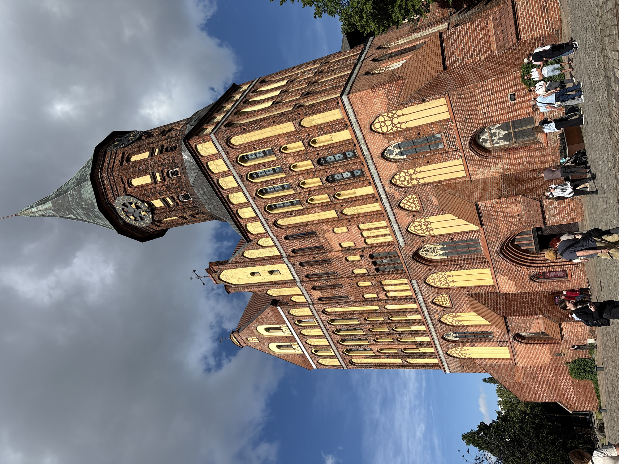 The image displays a large, historic brick building, likely the Kaliningrad Cathedral in Kaliningrad, Russia, captured under a partly cloudy sky with visible blue patches. The structure is predominantly reddish-brown brick, featuring a multitude of windows with distinctive pointed arches and intricate yellow or ochre-colored frames and tracery. A tall, dark-tiled spire, housing a clock tower, rises prominently on the left side of the building. The clock face, adorned with Roman numerals, shows the time as approximately 10:45 AM.
Several groups of people, casually dressed, are scattered on the paved ground surrounding the building, suggesting they are visitors or tourists. They appear to be observing the architecture and socializing in a relaxed manner. Green trees are visible along the periphery of the frame, hinting at a park or green area bordering the cathedral. The overall scene depicts a typical morning at a historical and architectural landmark, with no specific event occurring. The building's detailed brickwork, gothic architectural elements, and towering spire convey a sense of historical grandeur. Apart from the numerals on the clock face, no other legible text is visible.