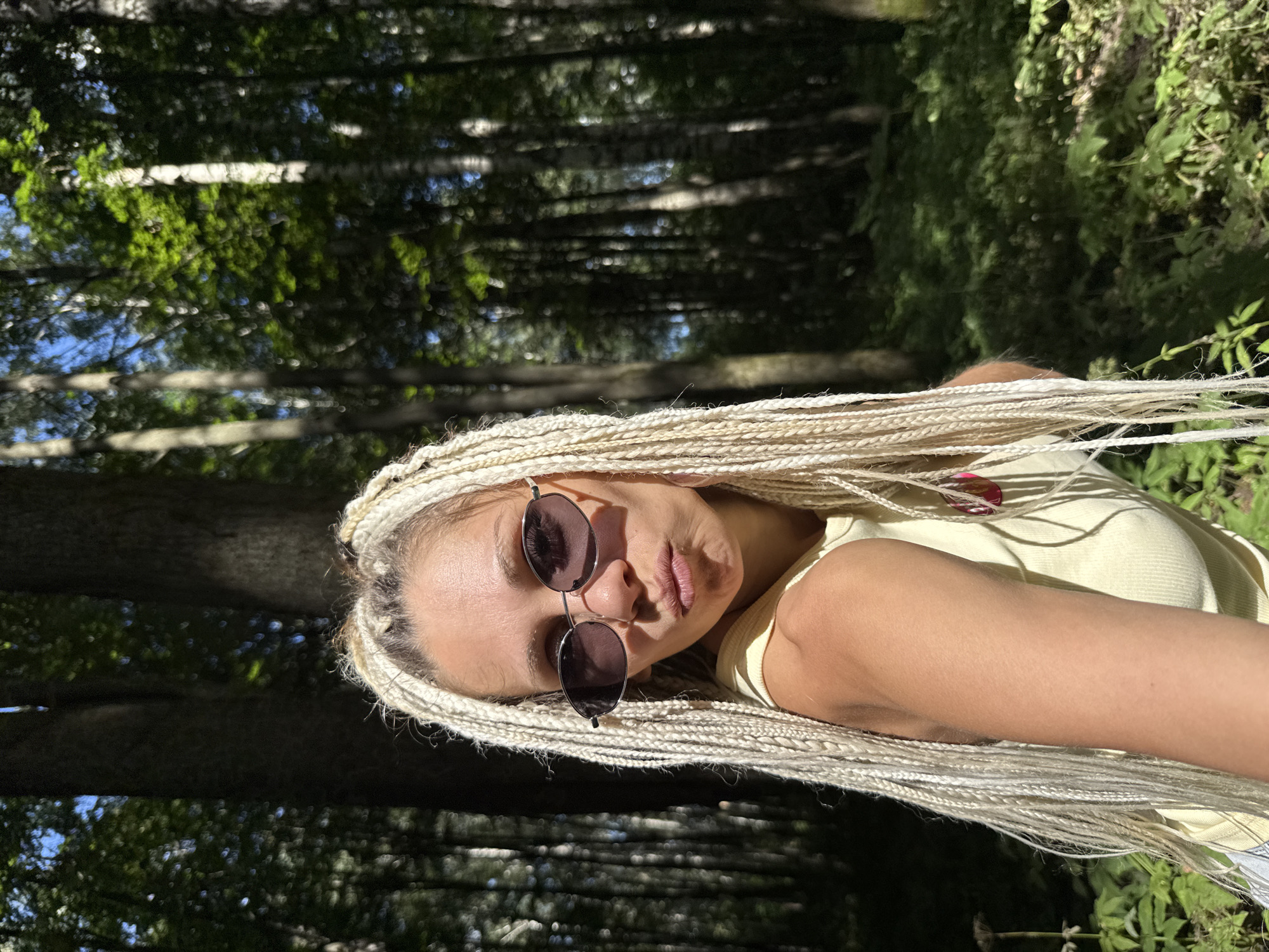 A young woman with blonde braids and sunglasses is taking a selfie outdoors. She is wearing a yellow tank top and has her lips pursed. The background shows a forest with tall trees and dappled sunlight. The location is specified as Rusanovo, Russia.