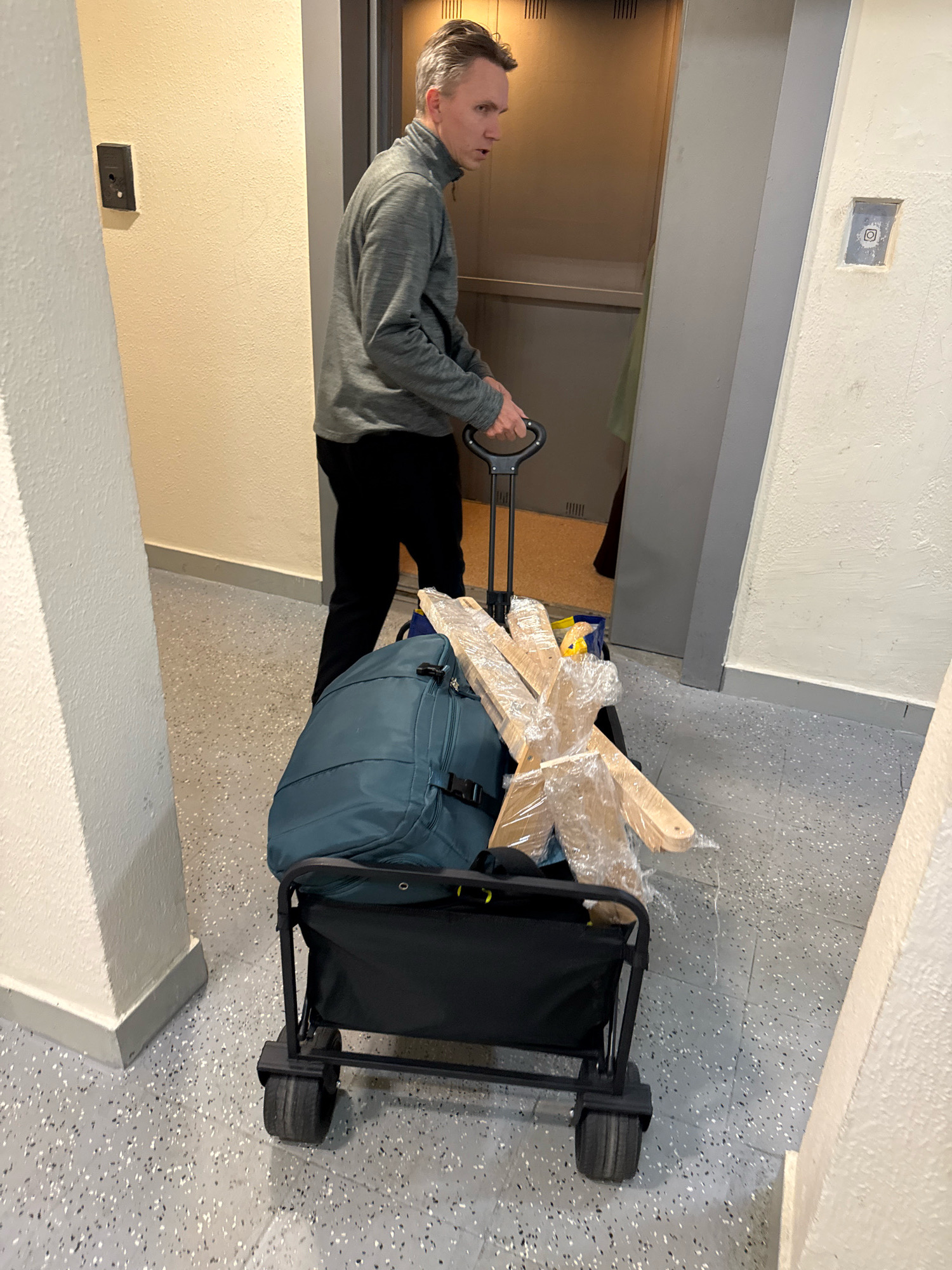 A man with short, light brown hair is standing in a hallway, pushing a black folding wagon. The wagon contains a teal duffel bag and several pieces of light-colored wood wrapped in clear plastic. The man is wearing a gray, quarter-zip sweatshirt and black pants. He is looking towards an open elevator door. The hallway has light yellow walls with a textured finish and a speckled gray floor. There are white pillars in the foreground. The scene appears to be in an apartment building.