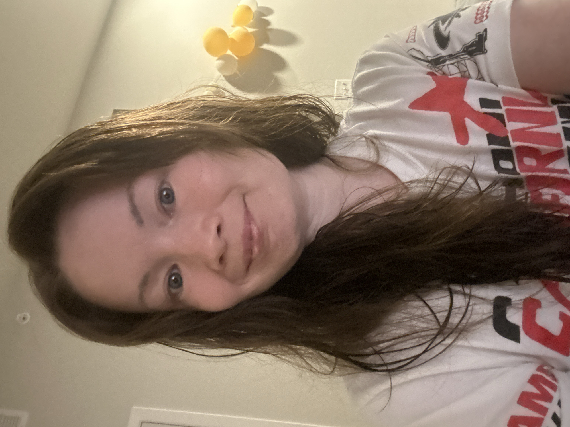 A young woman with long, dark brown hair and blue eyes is looking at the camera in a close-up selfie. She is wearing a white t-shirt with red and black graphics. The background is a light-colored wall with some yellow and white spherical objects attached to it, possibly a decorative item. There is also a light switch visible on the wall. The lighting appears to be indoor lighting. The image does not provide any information about the location in Colorado Springs, United States of America.