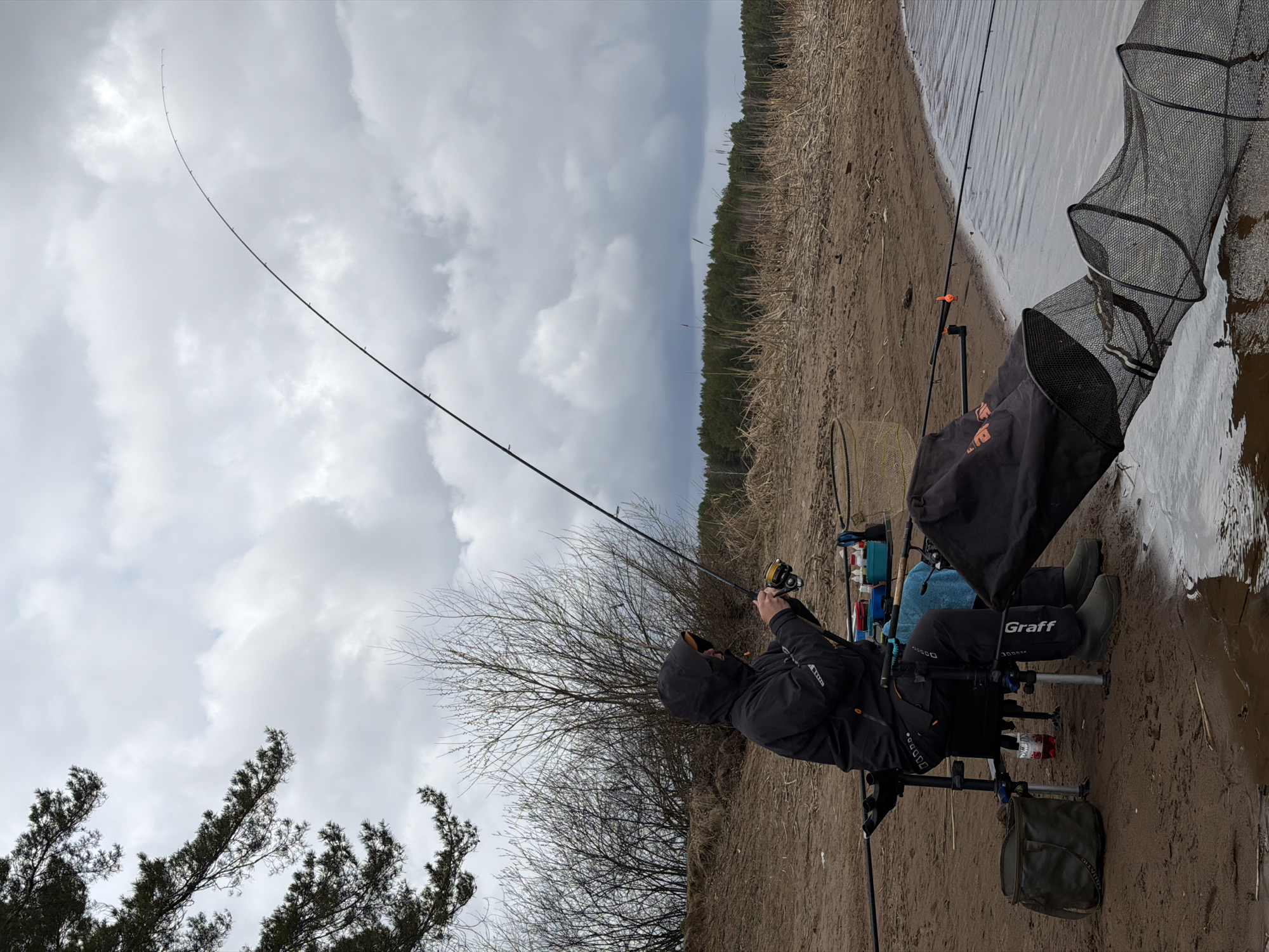 A man is fishing on the bank of a river or lake in Chaikovsky, Russia. The man is wearing a dark hooded jacket and dark pants, and is seated in a fishing chair. He is holding a fishing rod, which is bent with a fishing line extended. A landing net rests beside him. The environment includes a sandy bank, a body of water, and vegetation. The sky is overcast with cloudy formations.