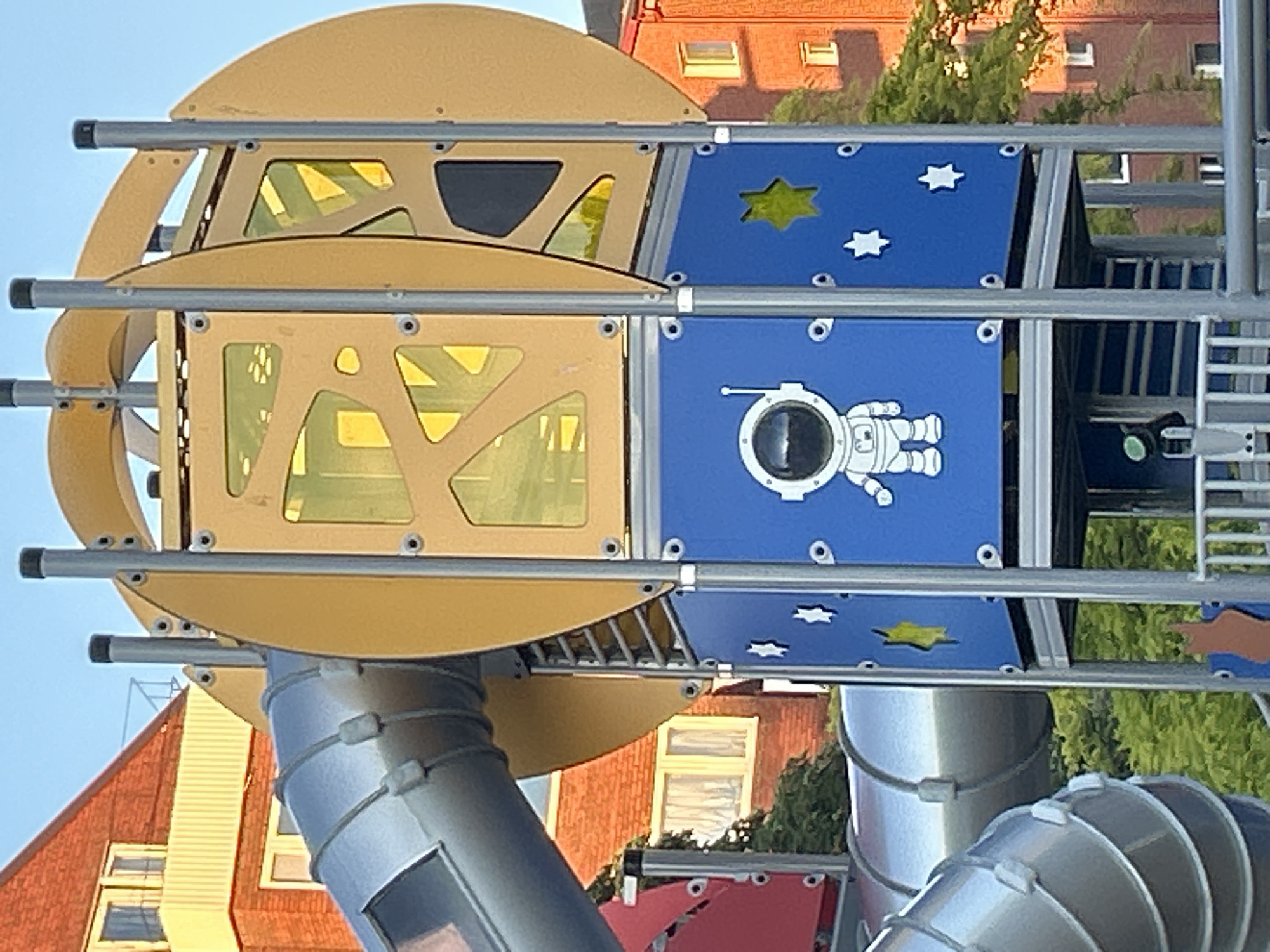 This is a close-up, eye-level, outdoor, daytime photograph of a section of a playground structure designed with a space theme. The structure is primarily composed of blue and yellow panels, with silver metal tubing forming slides and structural supports. A prominent blue panel features a cartoonish white astronaut graphic and several white and yellow star cutouts. Above the blue section, a yellow panel has intricate, abstract cutouts revealing yellow translucent material. Silver, corrugated tubing slides are visible to the left of the frame. In the background, a multi-story brick building with windows and balconies is partially visible. Lush green trees are also present. The overall impression is a bright, colorful, and playful playground element. This playground is located at Filbornaskolan in Helsingborg, Sweden.