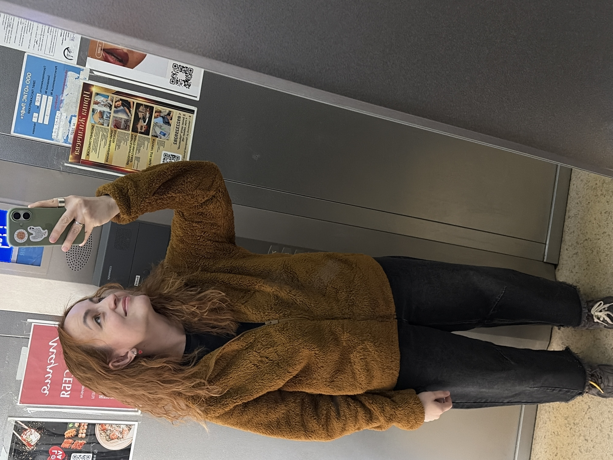 A woman with red hair stands in front of a mirrored wall. The wall has some posted advertisements, including one in Russian for sushi and another with the phone number +7 80553520585. There's also a blue sticker on the mirror, a white poster with a QR code, and other paper-based advertisements. The woman is wearing a brown fuzzy jacket and dark pants with worn, frayed edges, and appears to be taking a selfie with her smartphone. The floor appears to be made of stone or tile. The text on the advertisement is mostly in Russian, but also mentions the city of Chaikovsky.