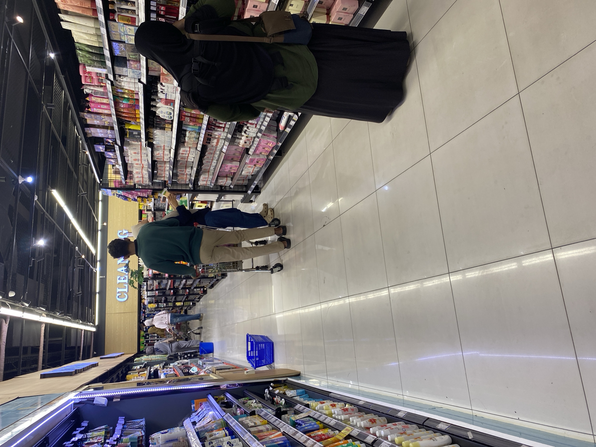 In a brightly lit indoor grocery store aisle, possibly in Banda Aceh, Indonesia, three individuals are visible. The person closest to the foreground, wearing a black hijab and an olive-green top and dark skirt, stands facing away from the camera, facing a shelf of products. To their left, a man in a green long-sleeved shirt and tan pants pushes a shopping cart. Further ahead and to the left, two more individuals, one in a light pink top and jeans and another in grey, are walking away from the camera. The aisle is lined with shelves stocked with various products, and the floor is tiled and appears clean. A blue shopping basket is placed on the floor in the middle of the aisle. The overall impression is a typical shopping scene in a modern retail environment.