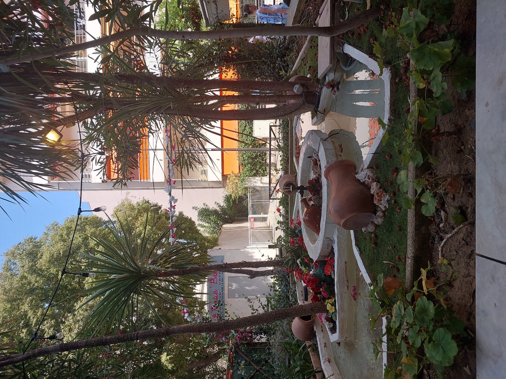 The image shows a courtyard with a small, tiered water feature in the foreground. The water feature is decorated with several large, terracotta amphorae and colorful flowers. Lush green foliage and palm trees surround the water feature, with a building in the background. A sign with French text, "Rose Bar", is visible on the building, suggesting a commercial establishment. In the distance, a person in a light blue dress is walking away from the camera. The environment appears to be an outdoor seating area or garden, possibly belonging to a restaurant or bar in Antibes, France.
