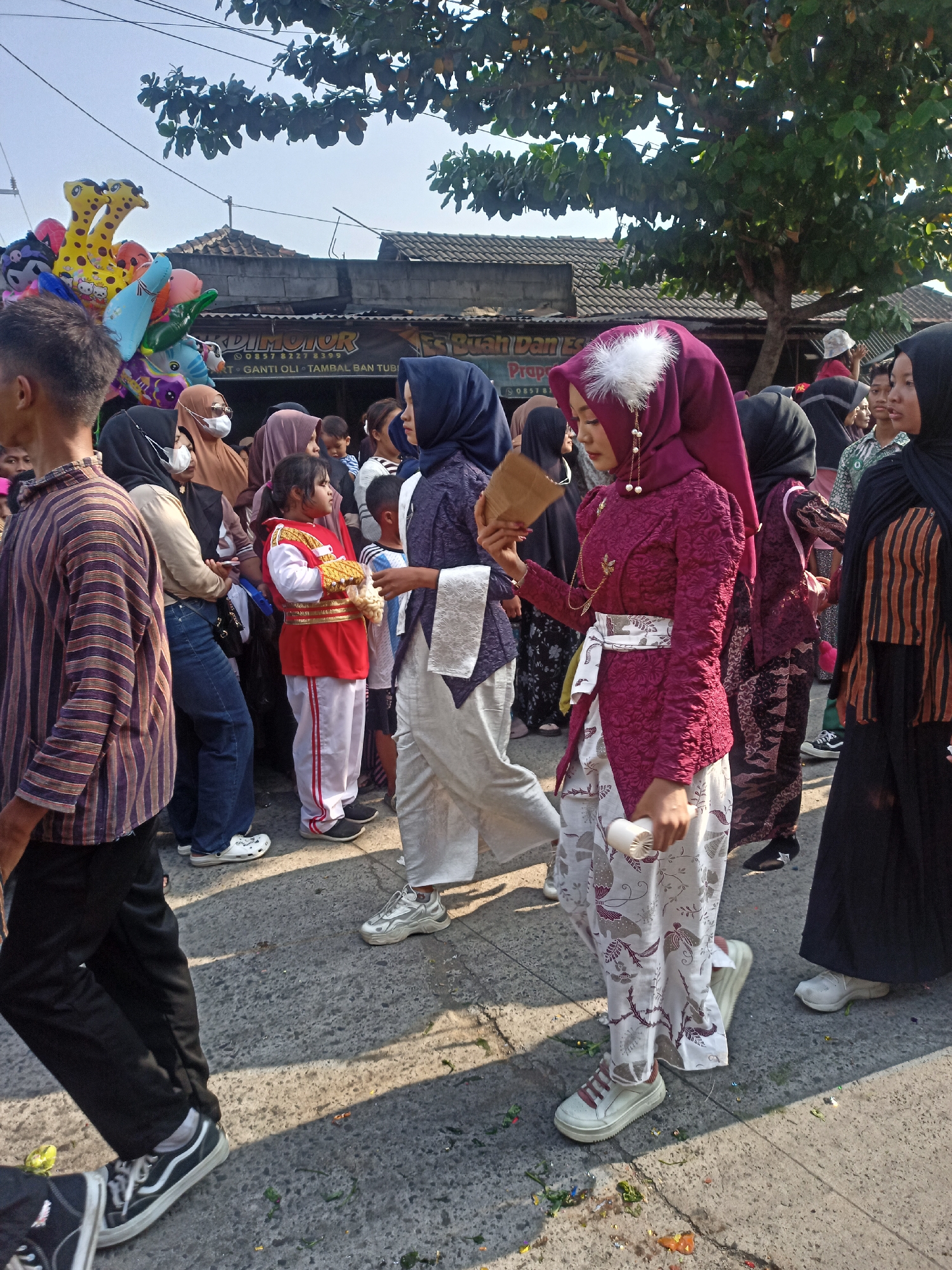 In Saren, Indonesia, on a paved street lined with buildings and trees, a group of people is gathered. The foreground shows the lower bodies of several individuals in motion, suggesting a procession or event. To the left, a person in a striped long-sleeved shirt and black pants walks towards the viewer. Next to them, a person in blue jeans and a beige shirt is partially visible. In the center, a young girl in a red and white uniform stands holding some items, facing right. She is surrounded by other people, some wearing hijabs and masks. To the right, a woman in a vibrant purple outfit, including a hijab and patterned skirt, walks towards the right. She is holding a small brown object and a white rolled-up item. Behind her, other individuals in traditional attire are also present, some wearing hijabs. In the background, a building with signs, including "ardi motor" and a phone number, is visible. Balloons, some in the shape of giraffes, are clustered in the upper left of the image. The ground is covered with debris, including small pieces of paper and leaves. The scene appears to be outdoors during the daytime with sunlight casting shadows.