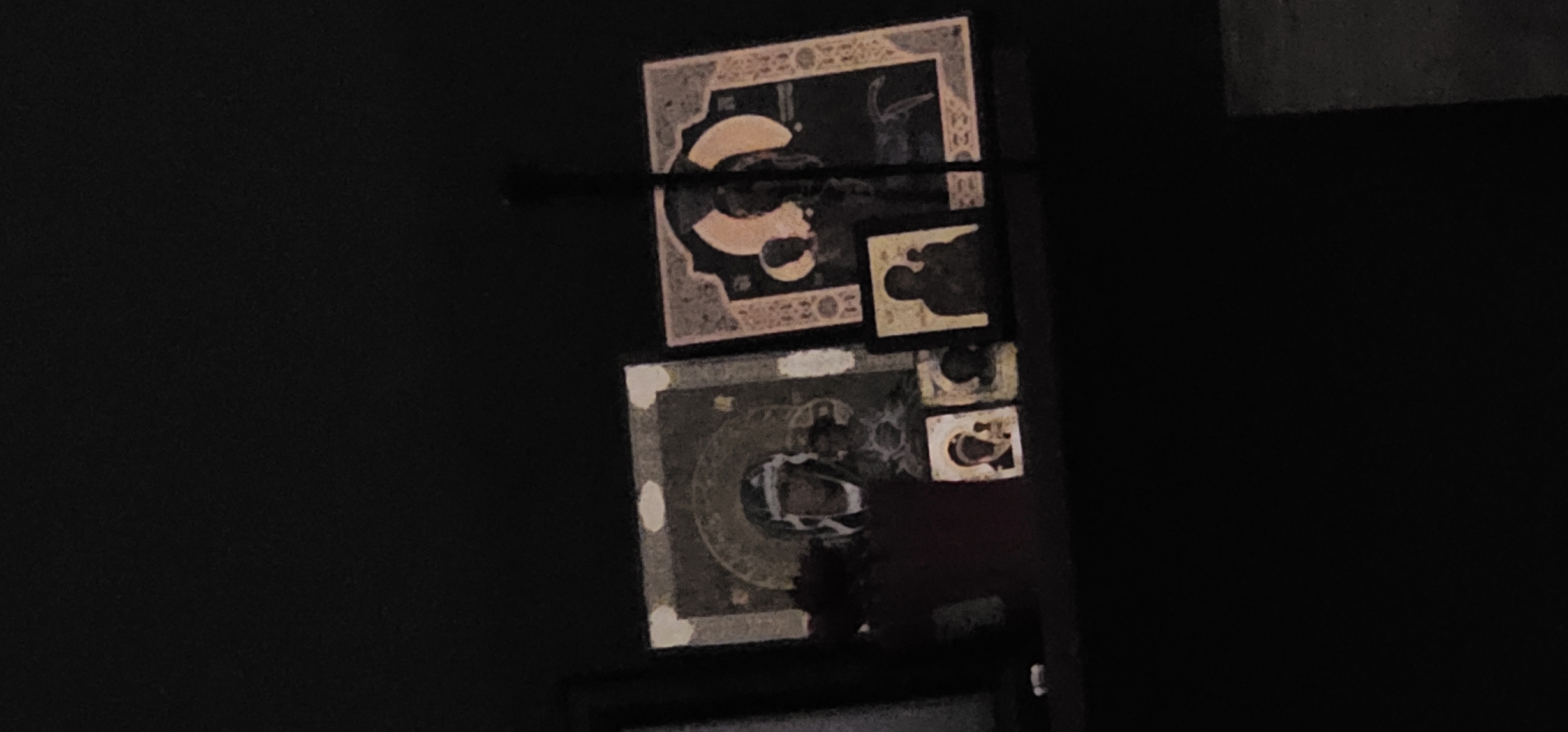 The image shows a collection of religious icons hanging on a wall in what appears to be a dimly lit interior space. The icons are arranged in a stacked manner, with a larger, square-shaped icon on top and another below it. The icons feature intricate designs and depictions of figures, typical of religious iconography. The lighting is poor, making it difficult to discern fine details. The location within the city of Unknown, Unknown, is not discernible from the image.