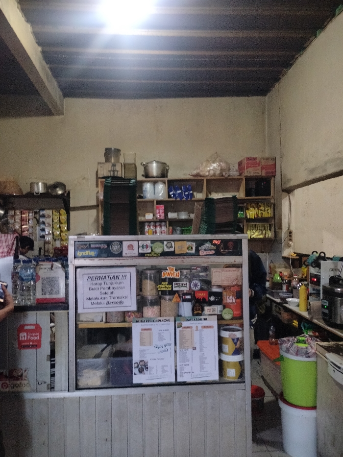 This image captures the interior of a small, informal eatery or warung, likely located in Dramaga, Indonesia. The setting appears functional, featuring a central glass-fronted counter that serves as a display and transaction point, surrounded by shelves stocked with various goods.
On the left, a partially visible person holds a phone, positioned near a rack holding packaged drinks and prominent QR code payment options for ShopeeFood, GoPay, and GoFood. The main counter displays jars of ingredients or toppings and is adorned with various stickers. A significant sign on the counter, in Indonesian, reads: "PERHATIAN !!! Harap Tunjukkan Bukti Pembayaran Setelah Melakukan Transaksi Melalui Barcode" (ATTENTION!!! Please Show Payment Proof After Transacting Via Barcode), outlining the payment procedure. Two menu lists, "DAFTAR MENU PANCONG" (Pancakes Menu List) and "DAFTAR MENU" (Menu List), are also visible, along with an "Enjoy your meals" message and a "HALAL" sticker.
Behind the counter, wooden shelves are neatly arranged with various supplies including instant noodles, packaged snacks, plastic-wrapped items, and cooking pots. To the right, a preparation area is visible with kitchen appliances such as a blender, a rice cooker, and a yellow condiment dispenser. Another person, likely staff, is partially obscured behind the counter in this area, engaged in activity.
The space is illuminated by bright overhead artificial lighting, suggesting it is either evening or night, or simply an interior with limited natural light. The ceiling features exposed wooden beams, and the walls are plain, contributing to the casual ambiance of the local establishment. Visible brand names include "Agnamus," "THE BANVELI," and "GETR."