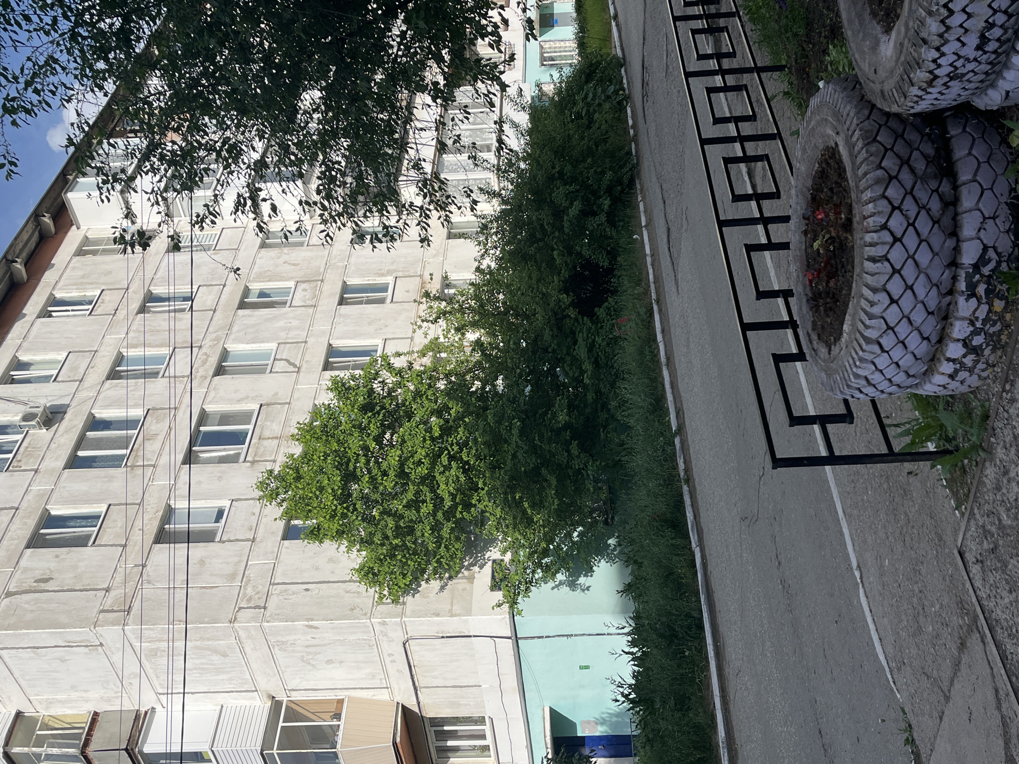 An outdoor shot of an apartment building in Chaikovsky, Russia. The building is made of light-colored concrete with rows of rectangular windows. There are trees partially obscuring the building, and a black metal fence runs along the side of a grey sidewalk. Stacked tires, painted white, with flower gardens inside, are also visible. Power lines run up the side of the building.