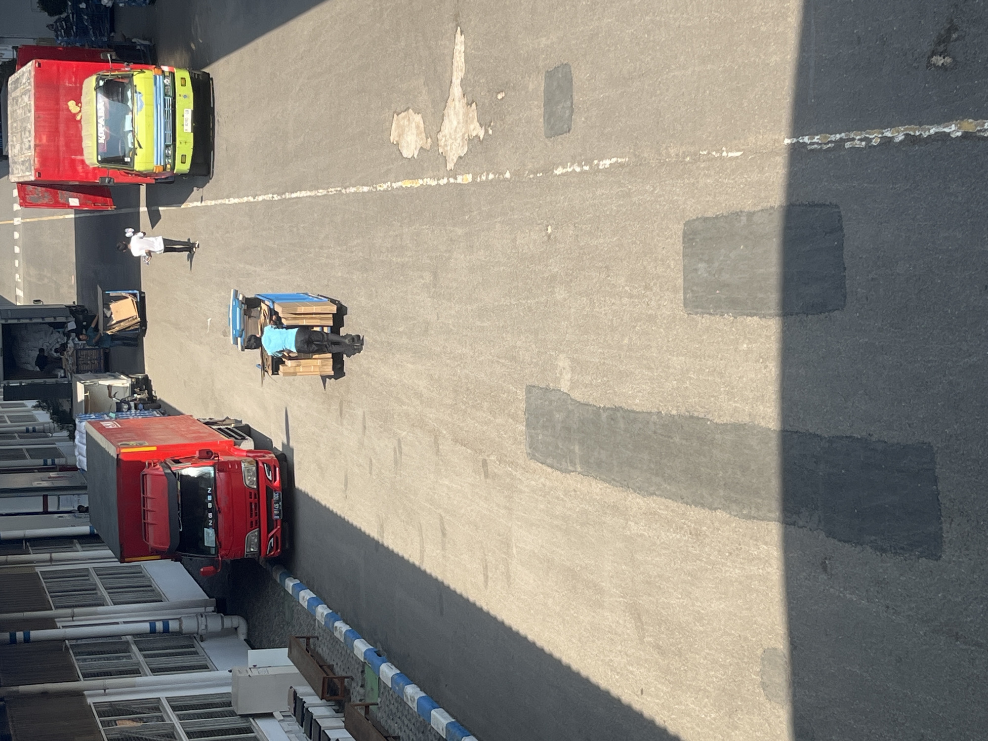 This is a high-angle, medium-shot photograph taken outdoors in Pelang, Indonesia, on a sunny day. The scene depicts a paved area, likely a loading dock or industrial yard, with several vehicles and individuals engaged in activity.
In the foreground, a person wearing a blue shirt and black pants is pushing a blue cart loaded with cardboard boxes. The cart has a metal frame and is being maneuvered on the asphalt. Further down the paved area, a person in a white shirt and black pants is walking away from the camera. To the right, a yellow and red truck with its rear doors open is parked. The truck has "KOPATRA" written on its windshield. On the left side of the frame, a red truck is parked, with "NNG SSN" visible on its windshield. The buildings in the background appear to be industrial or warehouse structures with large windows. The ground is marked with a white dashed line. Shadows from the buildings and a large object are cast across the asphalt, indicating the direction of the sun. There are some lighter patches on the asphalt, possibly from oil or water stains.
