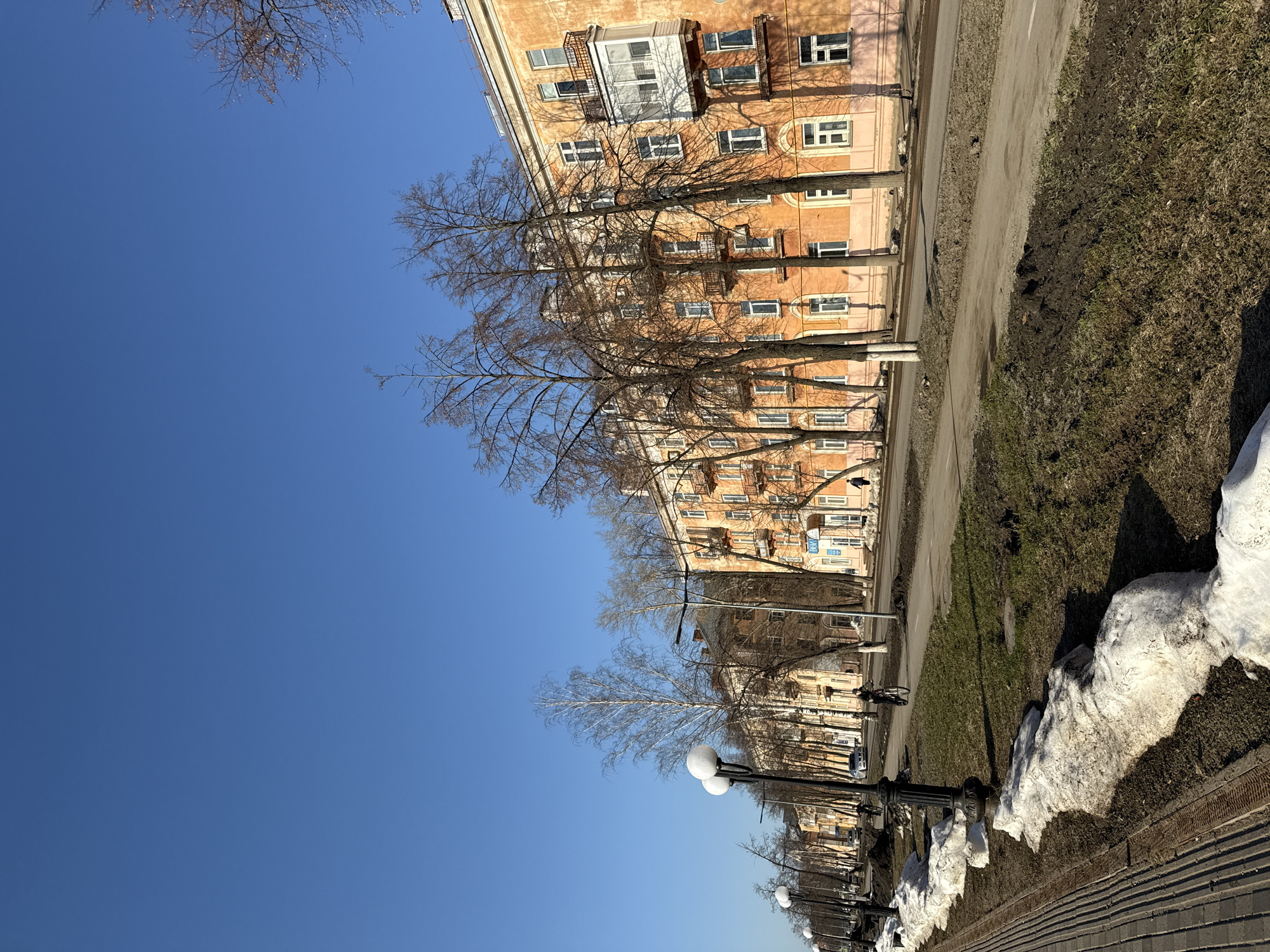 The image shows a street scene in Votkinsk, Russia, on a clear day. The sky is a solid blue. On the right side of the frame, a row of orange apartment buildings lines the street. Trees with bare branches stand in front of the buildings. Between the buildings and the sidewalk, there is a strip of land that is a mix of brown grass and melting snow. A road runs parallel to the buildings, and a sidewalk borders the road. A row of ornate, black lamp posts lines the sidewalk. The sidewalk is made of gray bricks. There are people walking along the sidewalk in the distance. Patches of snow are still visible on the ground.