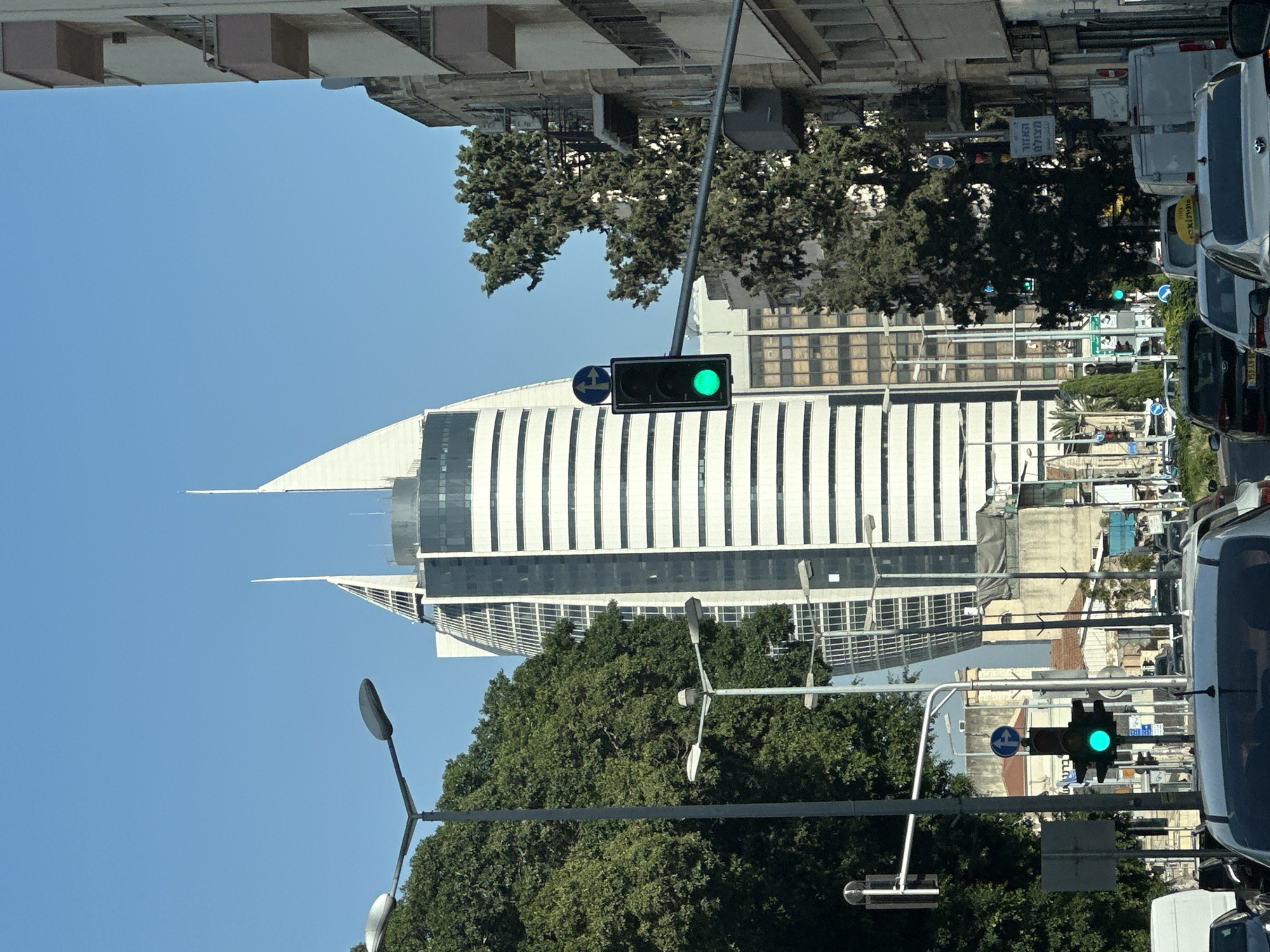 The image depicts a street scene in Haifa, Israel, characterized by a modern architectural style. Dominating the background is a tall, distinctive white skyscraper with a curved, sail-like facade, likely the "Sail Tower" (Bait Al-Malo'ah). The skyscraper features horizontal white bands and glass elements. In front of it, a lighter-colored building with horizontal white stripes is visible. To the right, an older, multi-story building with balconies and a beige facade stands.
In the foreground, several streetlights and traffic signals are present. One traffic signal displays a green light. Trees with dense green foliage line the street, contrasting with the urban structures. The road is populated with vehicles, including cars and vans, indicating traffic. The sky is clear and blue. The overall impression is a blend of historical and contemporary urban development.