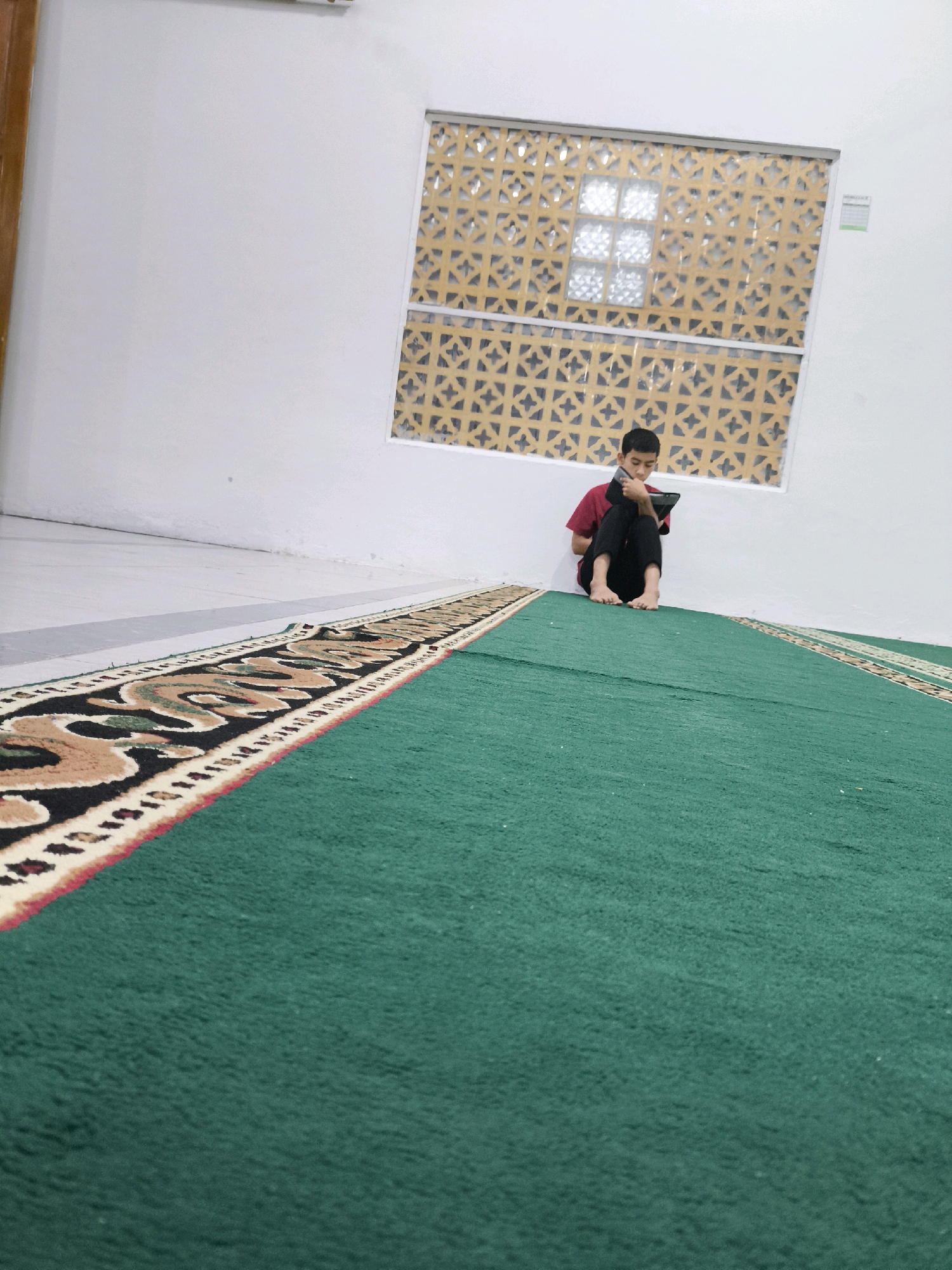 An Indonesian boy is sitting on the floor with his back against a white wall, reading on a tablet. He is wearing a red shirt and black pants, and is barefoot. The room has a large green carpet with a patterned border. There is a decorative window above him, with yellow and brown geometric patterns. The setting appears to be indoors, possibly a mosque or community center in an unspecified Indonesian city.