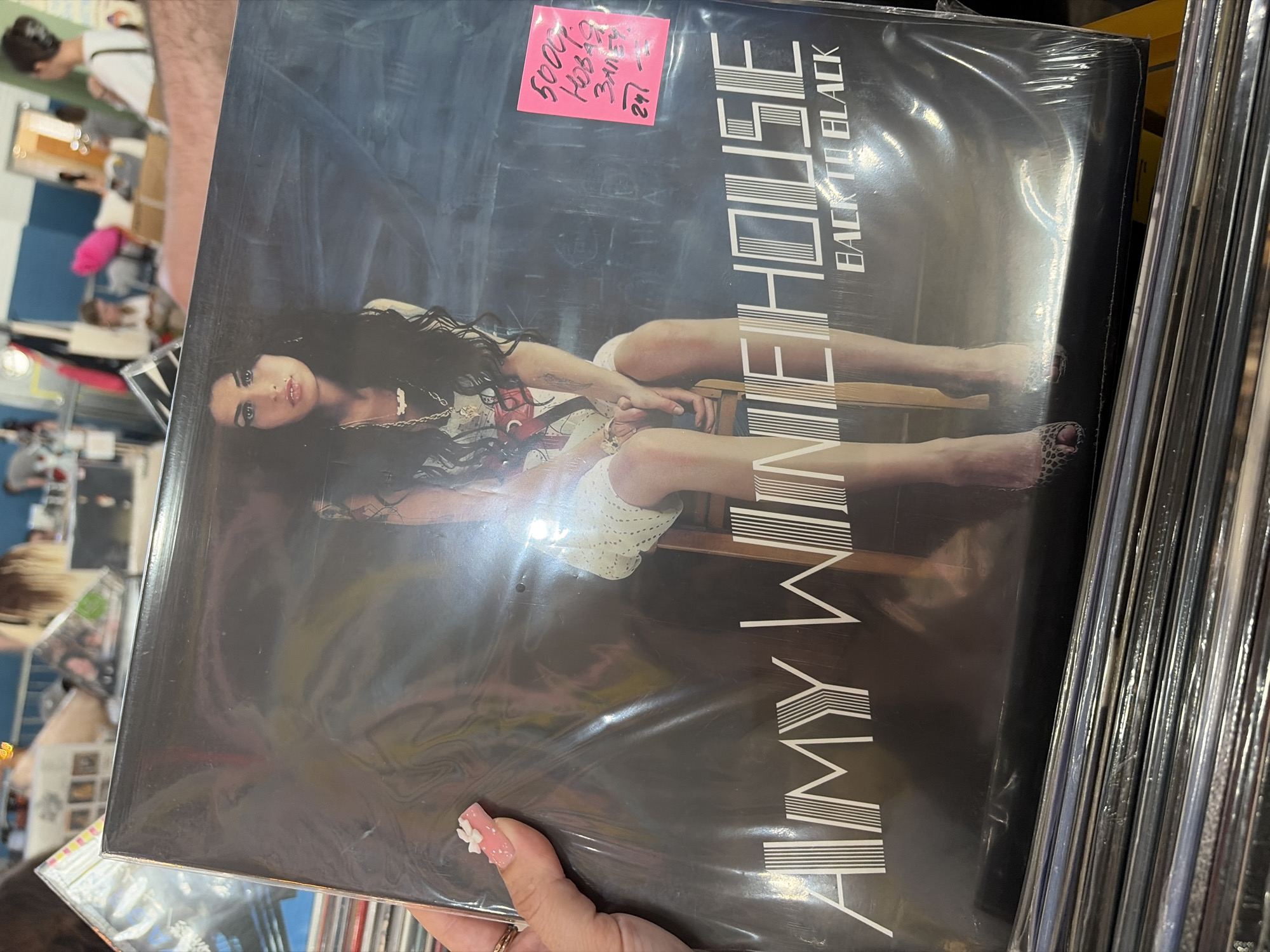 A close-up, eye-level shot captures a left hand holding a sealed vinyl record of Amy Winehouse's album "Back to Black" in an indoor commercial setting, identified as Saint Petersburg, Russia, due to Cyrillic text on a price tag. The hand, adorned with a manicured pink nail featuring a white bow embellishment and a gold-toned ring on the ring finger, positions the record prominently. The album cover displays Amy Winehouse seated on a wooden stool, looking directly forward, against a dark, textured background. Her name, "AMY WINEHOUSE," appears in a white, stylized font at the bottom left, with "BACK TO BLACK" positioned below it on the right. A rectangular pink sticky note is attached to the upper right corner of the clear plastic record sleeve. Handwritten in black Cyrillic, the note indicates "5000p," "Новая" (New), "Запись" (Record/Recording), and "24-". Below the held record, a stack of multiple other vinyl records, also appearing to be in protective sleeves, is visible horizontally. The background is blurred, showing several individuals in various positions, some seated and some standing, within a spacious indoor area with light-colored walls and distant blue wall sections, consistent with a record store or market.