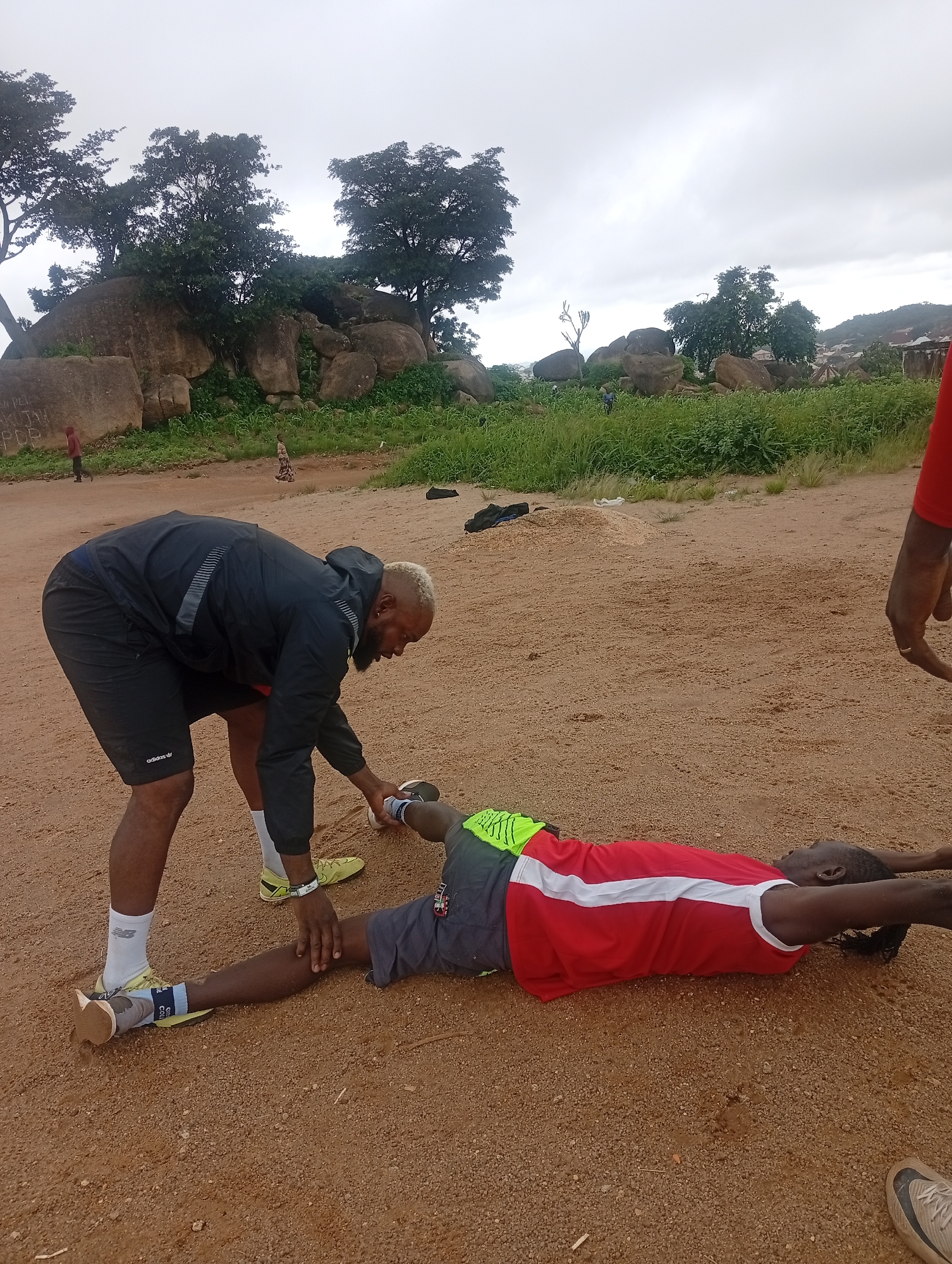 Two men are on a sandy surface. One man, wearing a black jacket and shorts, is bending over and holding the leg of the second man. The second man is lying on his back with his arms extended above his head, wearing a red shirt with a white stripe and dark shorts with green accents. They appear to be engaged in a stretching or physical therapy session. The background shows a landscape with large rock formations, trees, and vegetation, characteristic of the rocky terrain found in the city of Jos, Nigeria. The sky is overcast.