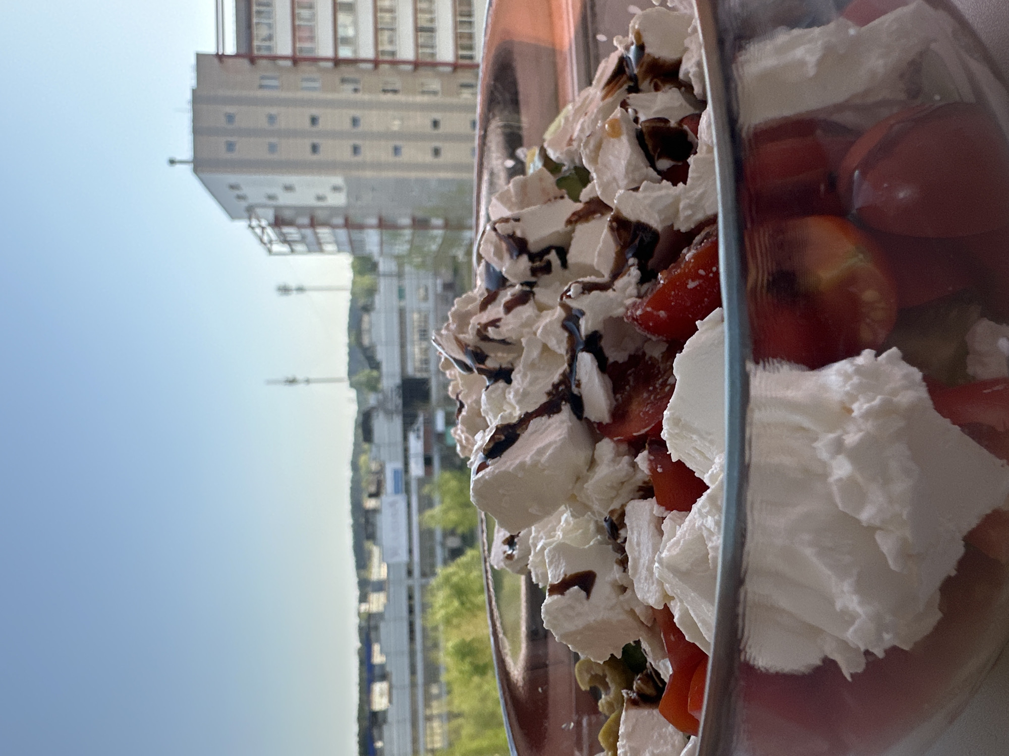 The image depicts a salad in a clear bowl, likely photographed on a balcony or near a window. The salad contains red tomatoes, white cheese cubes drizzled with a dark sauce, and other unidentified ingredients. Outside the window, an urban landscape is visible, including a tall apartment building with a repetitive window pattern and a smaller structure with a tower. The sky is clear and blue. Based on the location, the building in the background may be located in the city of Chaikovsky, Russia, but this is not definitively verified.