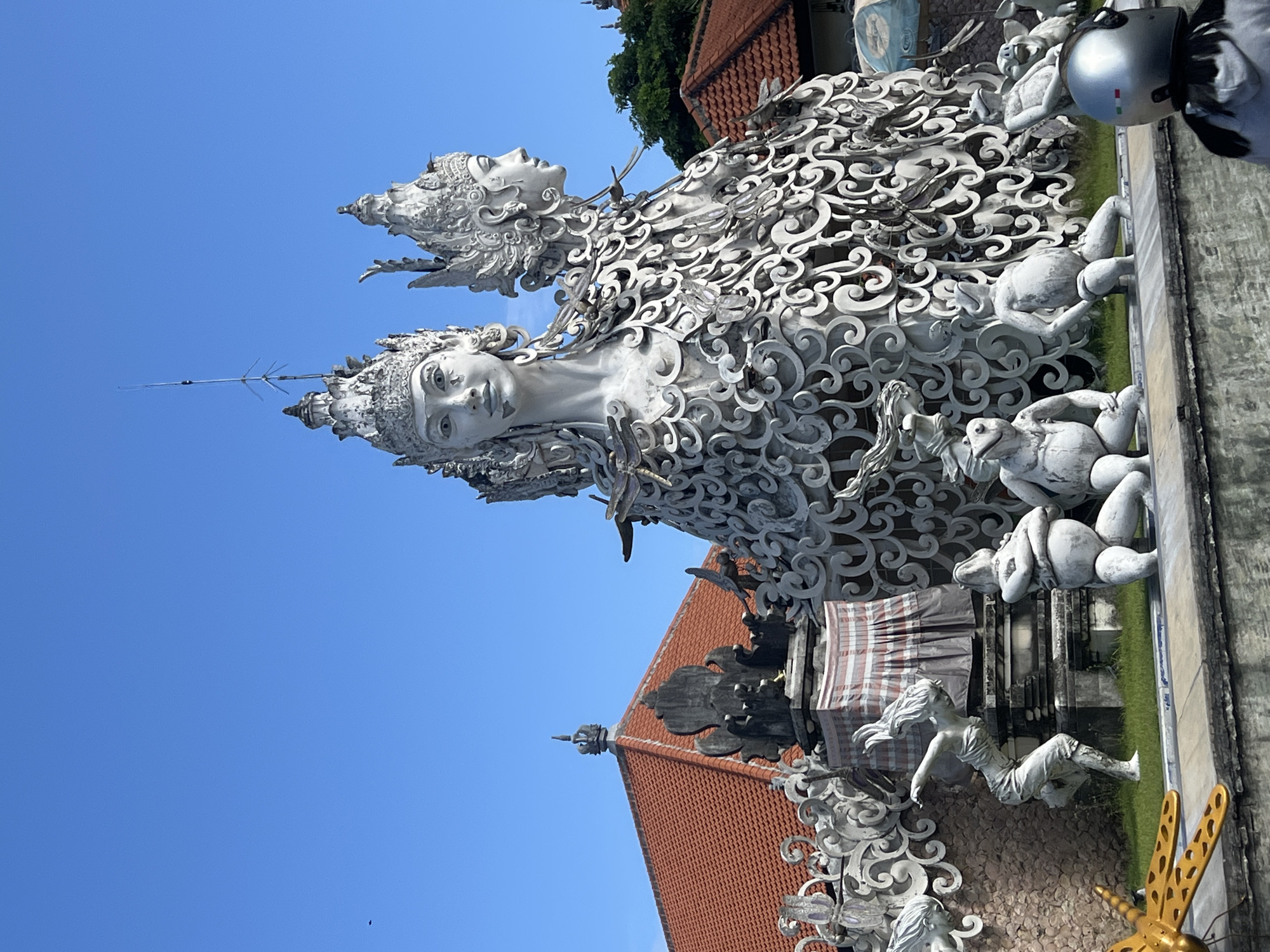 The image showcases a decorative statue complex in Sukawati, Indonesia, under a clear blue sky. Two large, white statues of female figures with ornate headdresses and intricate scrollwork forming their bodies are prominently displayed. Smaller white statues depicting human and animal forms are scattered around the base. A building with a red-tiled roof is visible behind the statues, and a person wearing a silver motorcycle helmet is partially visible on the upper right edge of the frame.