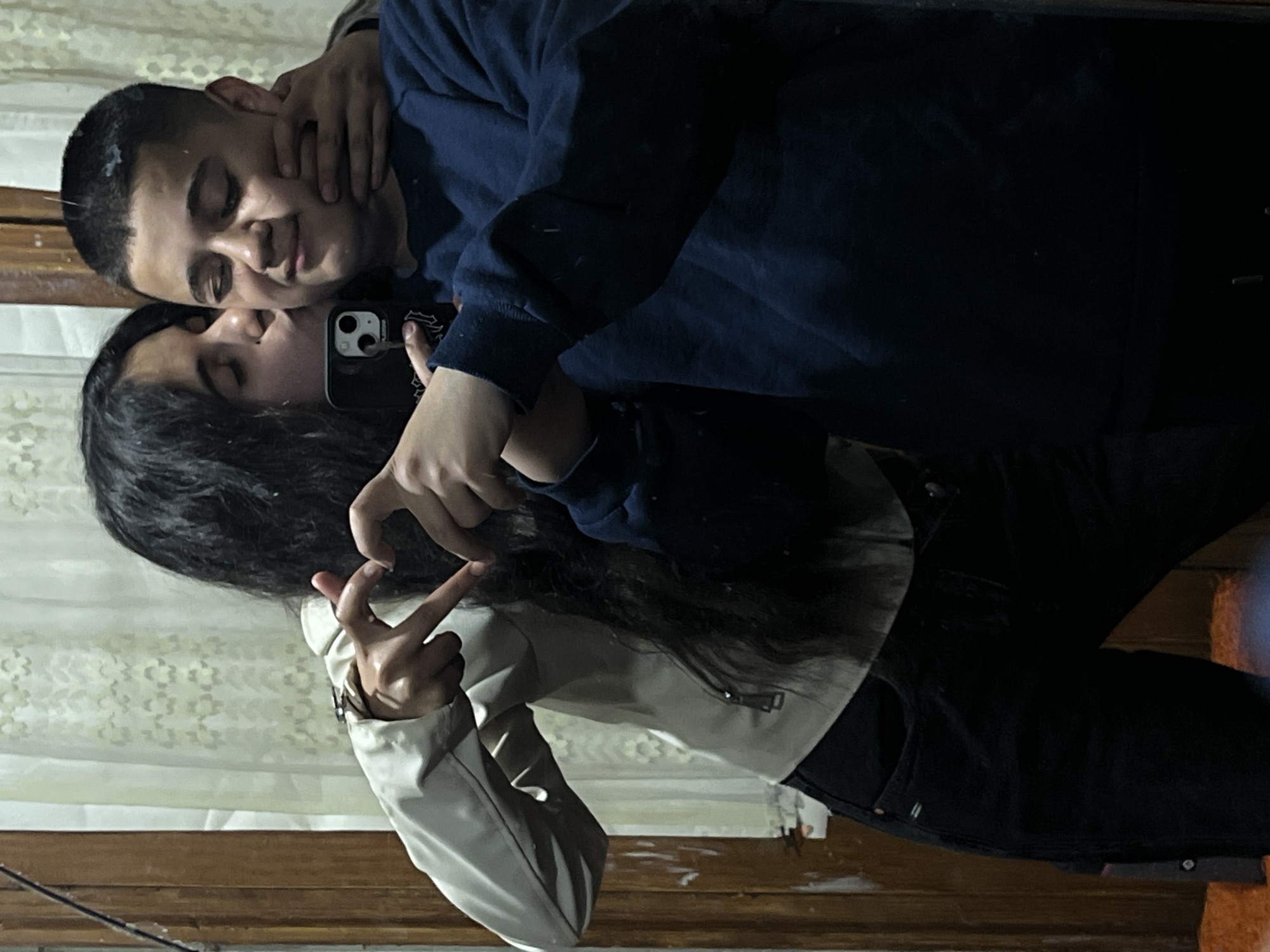 A selfie shows a young couple. The man, wearing a navy blue sweater, has his arm around the woman's neck. The woman, wearing a light tan jacket and black pants, is making a heart shape with her hands. They are standing indoors, likely in a home, as suggested by the wood paneling and curtained window in the background. The photo was possibly taken in Lozarevo, Bulgaria, based on the request. The man is holding a phone with an iPhone 13 Pro Max camera.