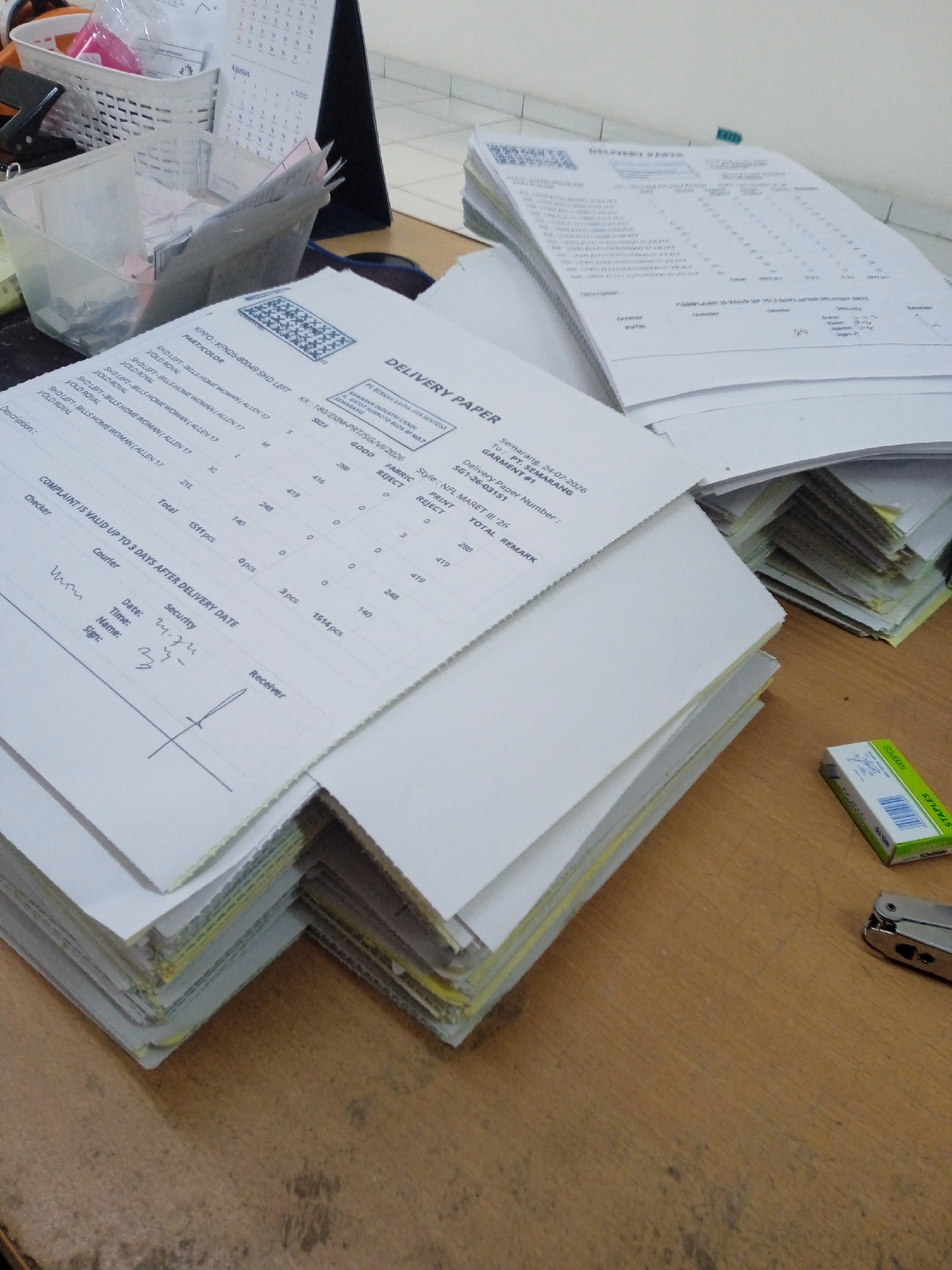 The image shows a desk with a stack of papers, a stapler, and a box of staples. The papers are delivery slips, and some of them contain information in Indonesian. The top paper has the words "DELIVERY PAPER" printed on it. There are also some handwritten notes and signatures on the papers. The box of staples is green and white and has the brand name "STAPLES" printed on it. The stapler is silver and appears to be a standard office stapler. The desk is made of wood and has a natural finish. The background of the image is out of focus but appears to be an office environment. The text on some of the papers indicates the location is Semarang, Indonesia.