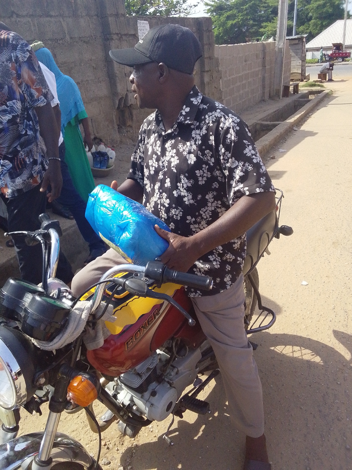 A man sits on a red and yellow "BOXER" brand motorcycle on a street in Lokoja, Nigeria. He wears a black baseball cap, a black and white floral print short-sleeve shirt, and tan pants. He holds a blue cylindrical package in both hands. Other people stand nearby, and a cinder block wall lines the street. The ground is sandy.