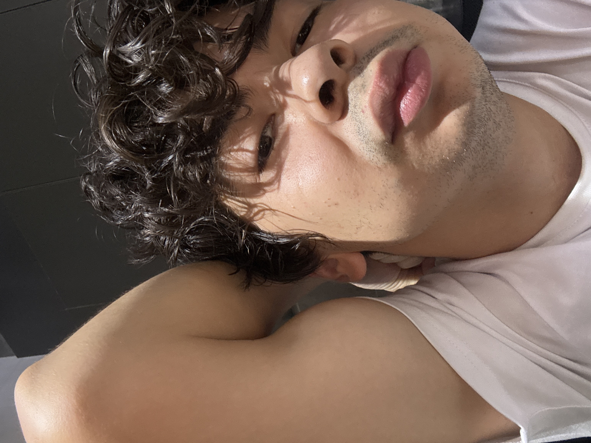 The image is a close-up, selfie-style shot of a young man with curly, dark hair. He is wearing a white t-shirt and is lying down, with his left arm bent behind his head. The lighting suggests an indoor environment, possibly a bedroom or living space, with natural light casting shadows on his face. There is no indication of a specific location in Tashkent, Uzbekistan, or any other city. The focus is on the man's facial expression, which appears to be a pout or grimace, and his physical features. The background is dark and indistinct, further emphasizing the subject.