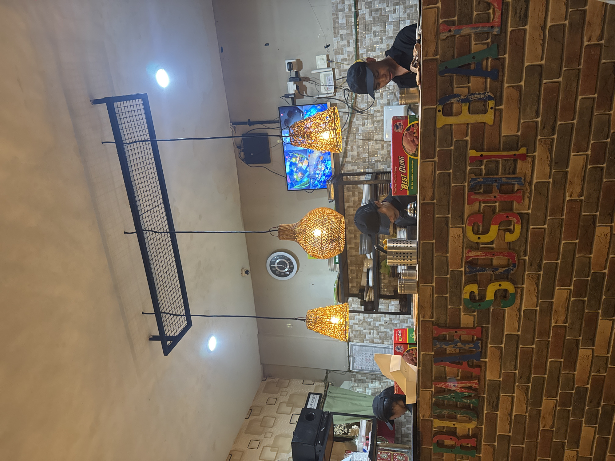 This media file captures an indoor scene, likely an eatery or convenience store, situated in Kuta Selatan, Indonesia. The image is oriented sideways, with the main focal point being a service counter on the right.
Two men, presumed to be staff, are visible behind the counter. One man, wearing a black cap, is positioned centrally and looks down towards the counter. Another man, also wearing a black cap, is partially visible at the far end of the counter, looking towards the camera.
The setting is illuminated by several artificial light sources, including bright overhead spotlights and three distinctive woven pendant lamps hanging at varying heights. A large flat-screen TV is mounted on the wall, displaying colorful animated graphics, possibly from a video game or cartoon. Above the TV, a small security camera (CCTV) is visible. A wall clock showing approximately 1:30 or 2:30 is also present.
The counter area features various items, including a "BEST CLING" brand plastic wrap box, which states "Quality Plastic Wrap" and "Cling Wrap Keeps food fresher and longer." To the left of the counter, a sign reads "STAFF ONLY." The background walls vary; one section features a pattern of square tiles, while the counter's front is adorned with a brick-patterned wall. Colorful, distressed wooden letters are affixed to the brick wall, partially spelling out words like "BEST" or other fragmented text that appears to be part of a larger sign. An open metal grid shelf is mounted on the wall to the left. The scene appears to be taking place at night or indoors where artificial lighting is necessary.