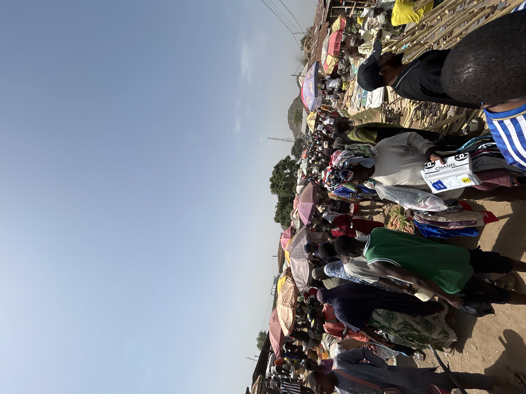 The image shows a crowded open-air market in Bukuru, Nigeria. People are shopping and selling items. Many colorful umbrellas provide shade. The ground appears to be dirt or packed earth. Merchandise is spread out on the ground. There is a mountain in the background. There are electricity poles in the background.