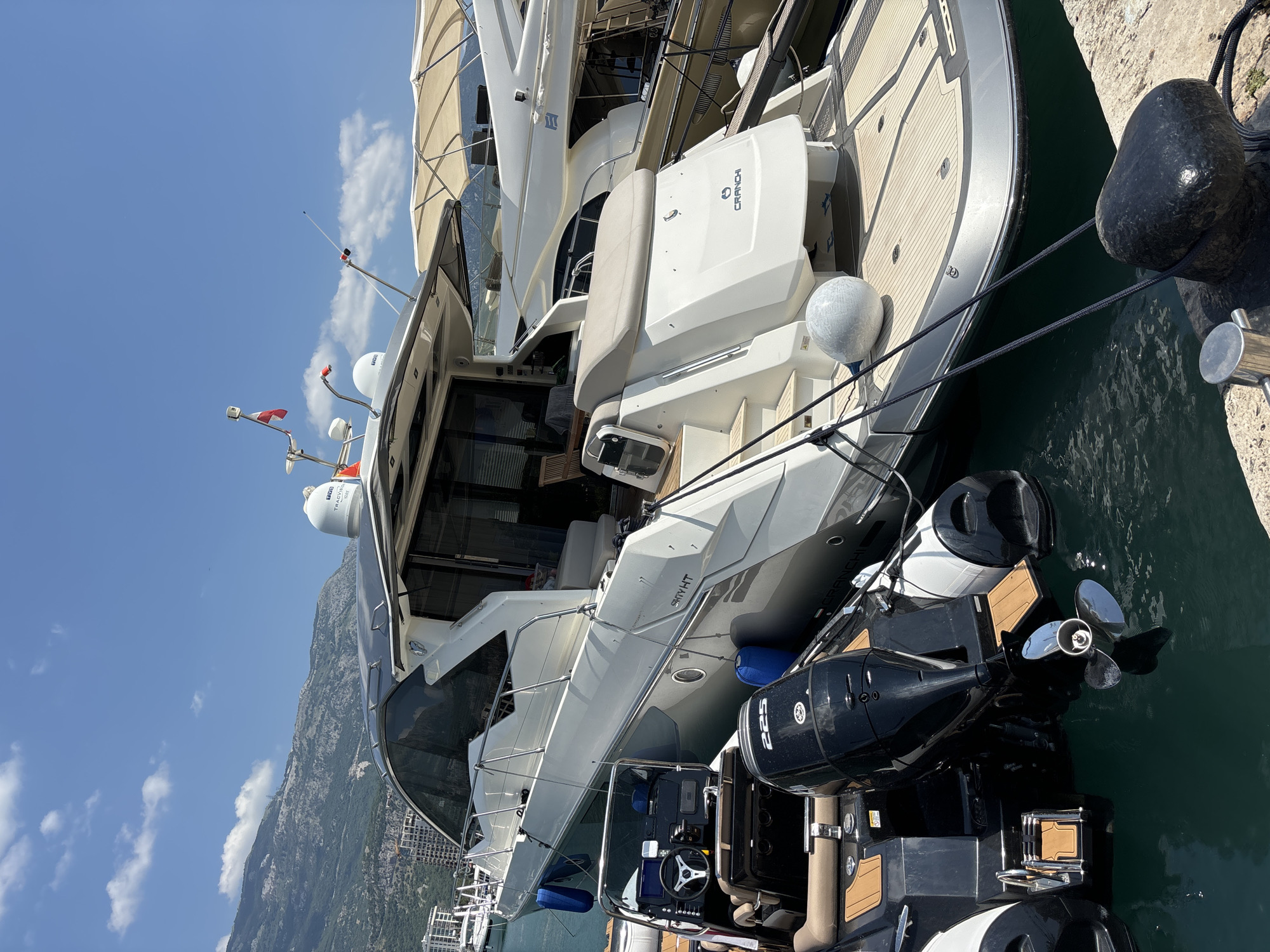The image shows two boats docked in the marina of Budva, Montenegro. The foreground is dominated by a black inflatable boat with two powerful outboard motors, one of which is partially submerged in the dark blue water. In the background, a large white yacht with a sleek design and dark windows is moored. The yacht has a wooden deck at the stern and the name "CRANCHI" is visible. The scene is set against a backdrop of a clear blue sky with a few scattered clouds, and a mountainous coastline with buildings can be seen in the distance. The overall impression is one of a sunny day at a luxury maritime destination.