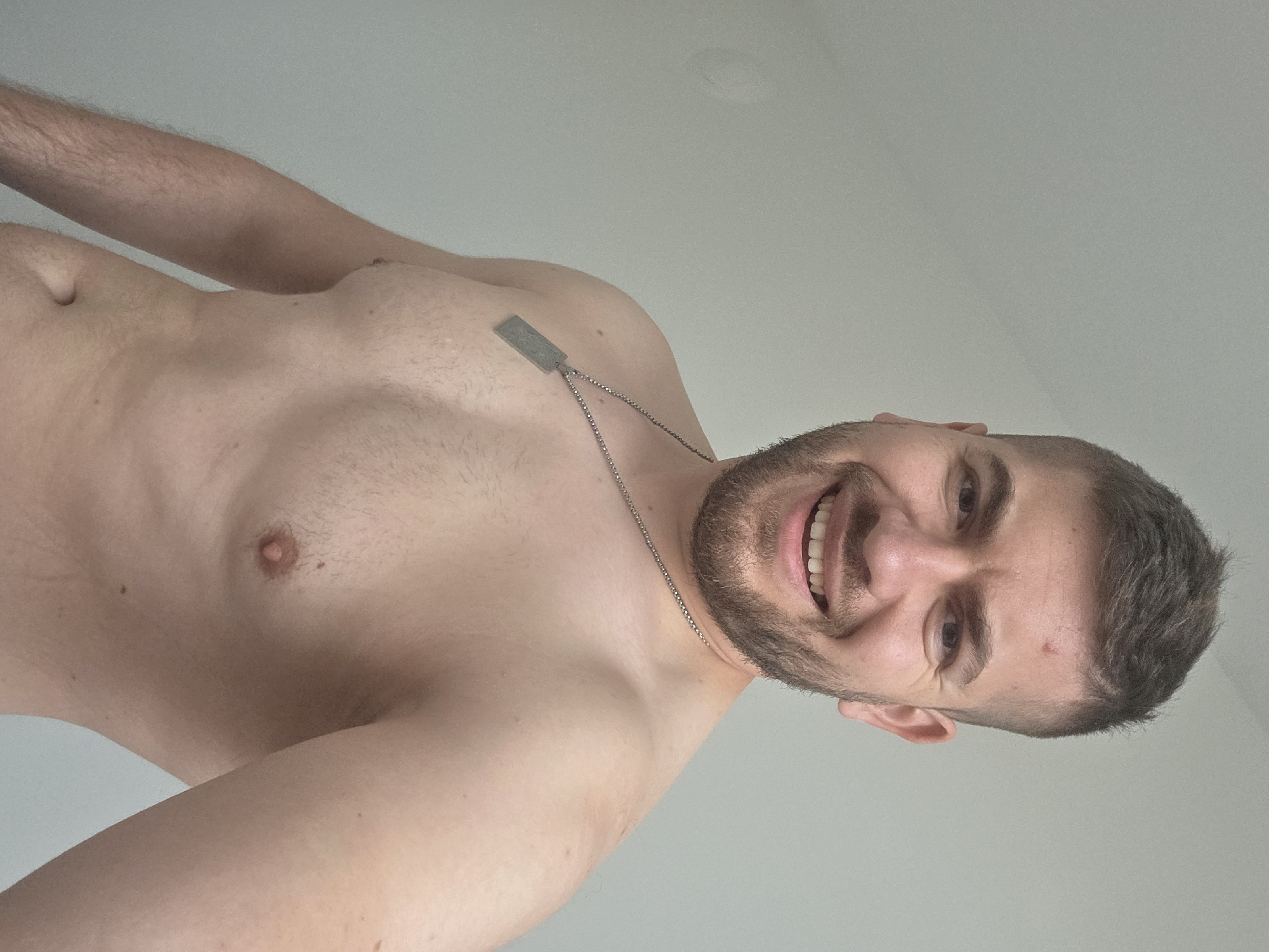 A shirtless man with short brown hair and a beard is smiling at the camera. He is wearing a silver necklace with a rectangular pendant. The background is a plain white wall. The location is not discernible from the image.