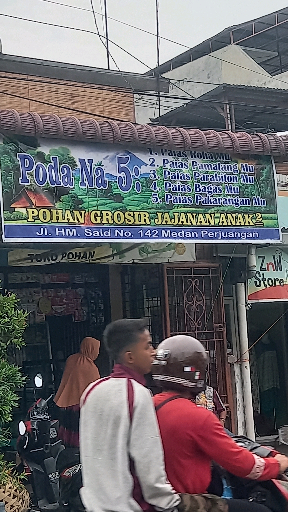 The image shows a street scene in the City of Medan, Indonesia. A prominent banner displays the text "Poda Na 5" and "POHAN GROSIR JAJANAN ANAK2" with an address "Jl. HM. Said No. 142 Medan Perjuangan". The banner is hanging from a building with a brown woven facade. Below the banner, a shop with the sign "TOKO POHAN" is visible. There are people and a motorcycle in the foreground. One person is wearing a white and maroon jacket, and another is wearing a red shirt and a brown helmet. Another person in the background is wearing a brown hijab. The environment appears to be an urban setting with a cloudy sky.